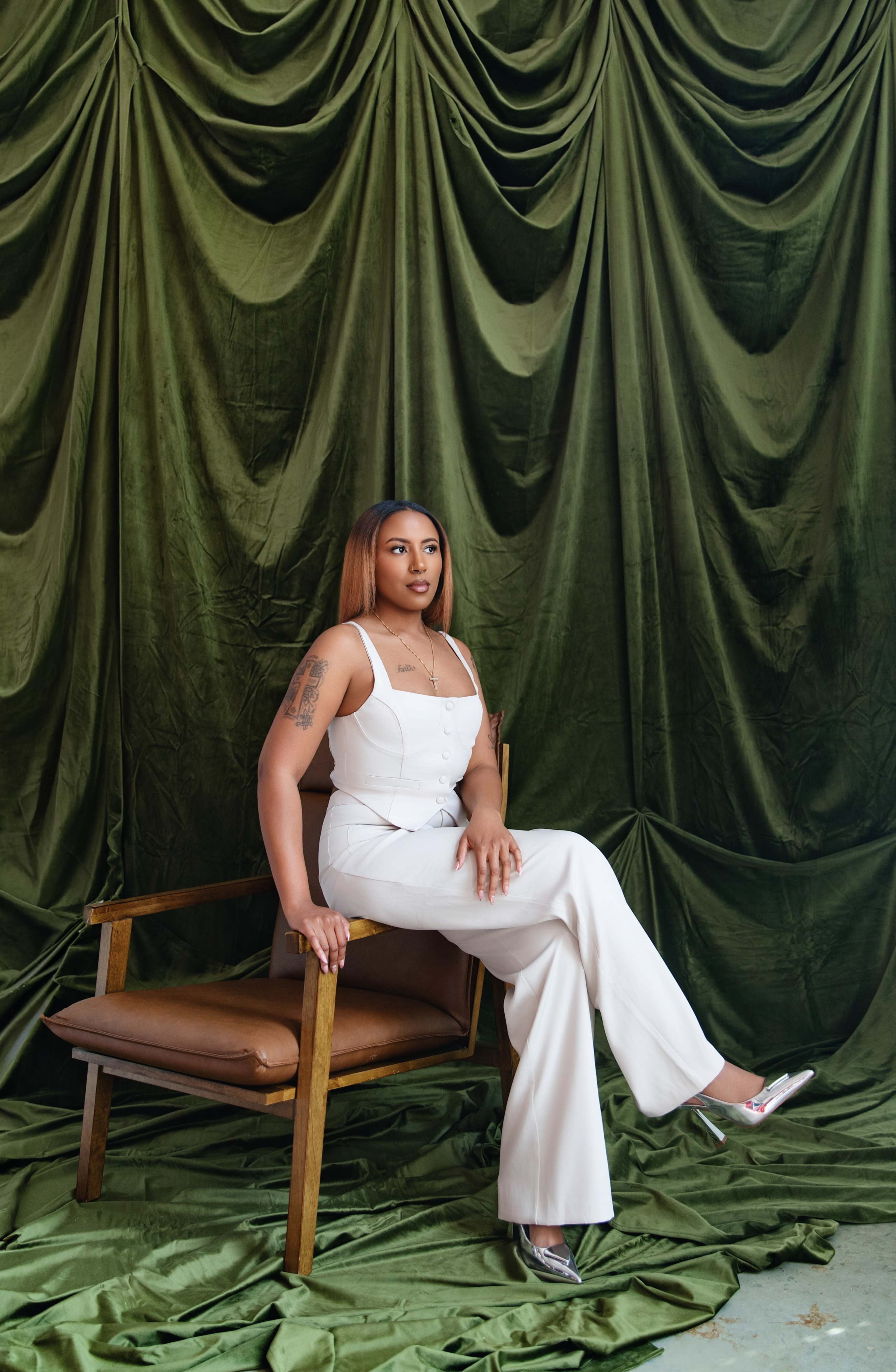 A woman in a white jumpsuit sits on a wooden chair against a green draped backdrop.