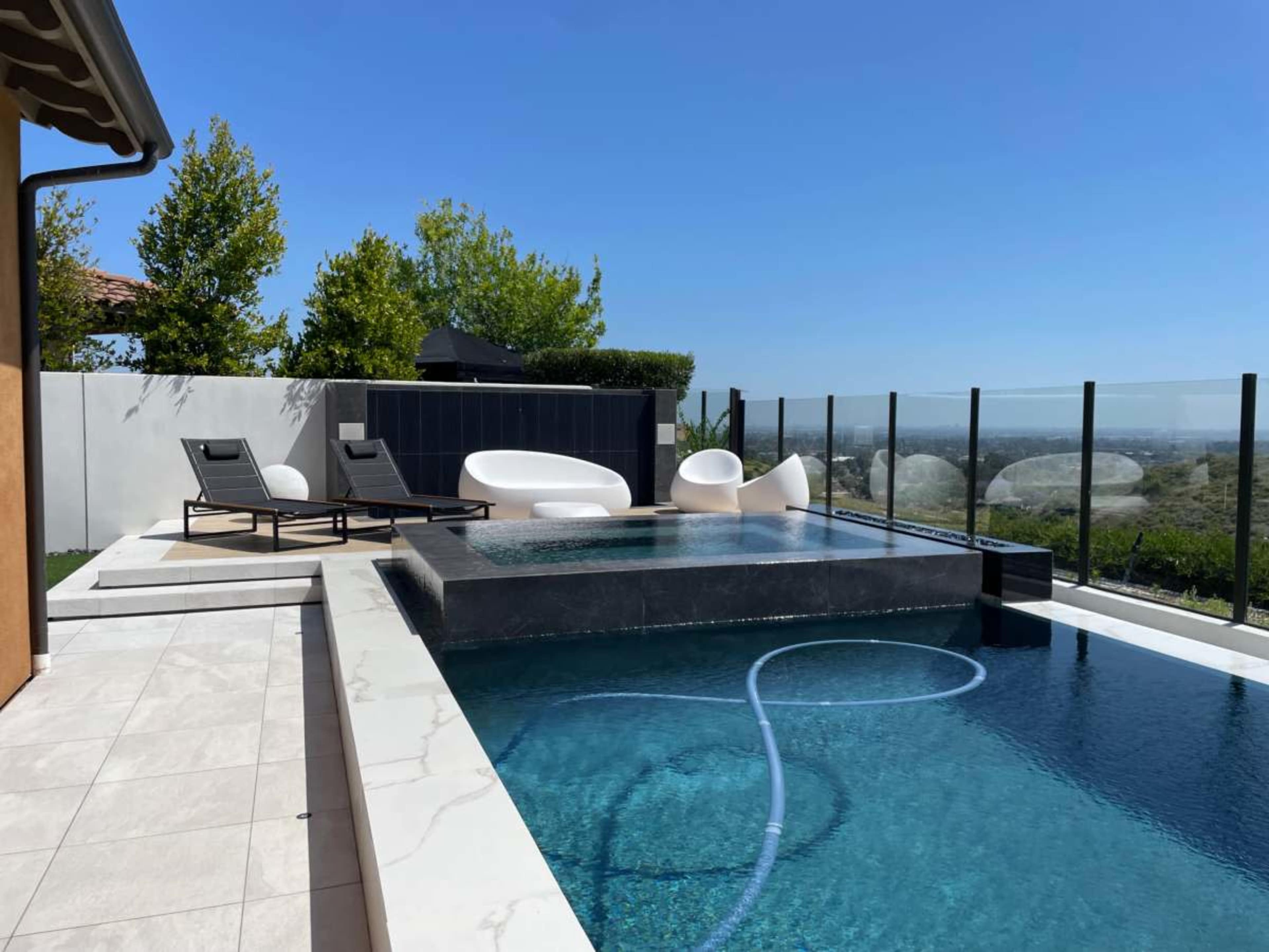 A modern pool area with a small infinity pool, lounge chairs, and a clear glass railing overlooking a scenic landscape.