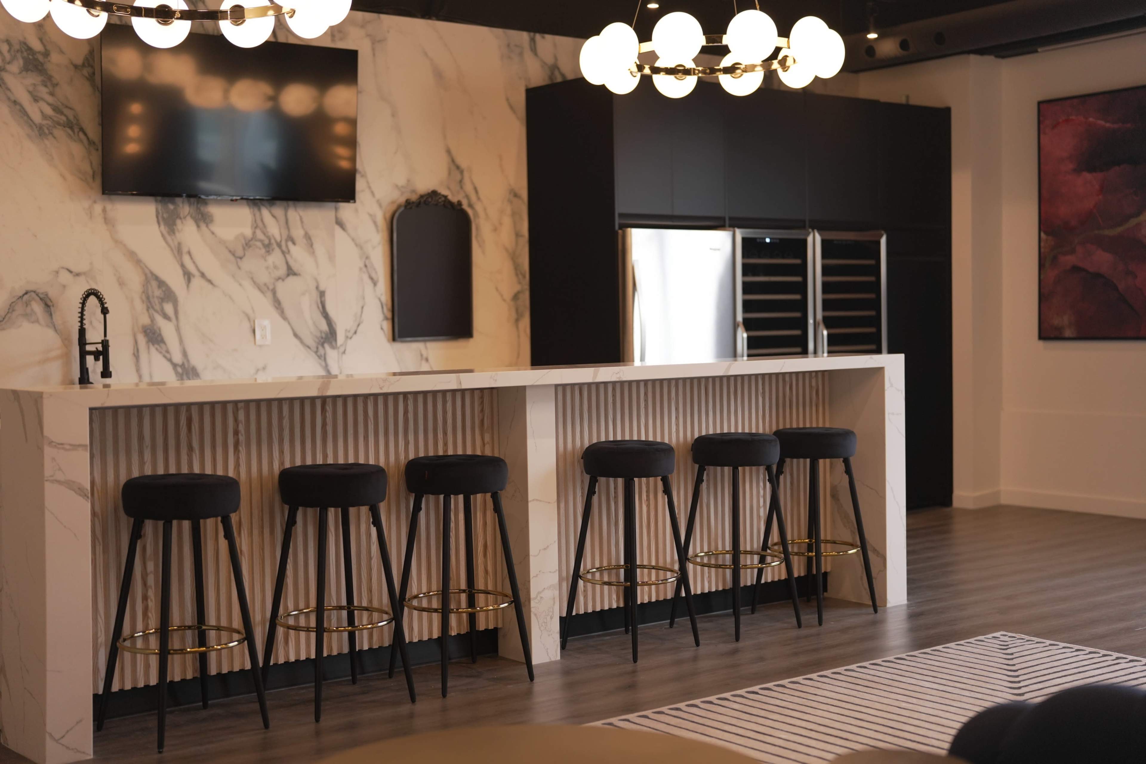 The image shows a modern kitchen with a marble countertop, black bar stools, and a refrigerator integrated into cabinetry, complemented by stylish lighting fixtures.