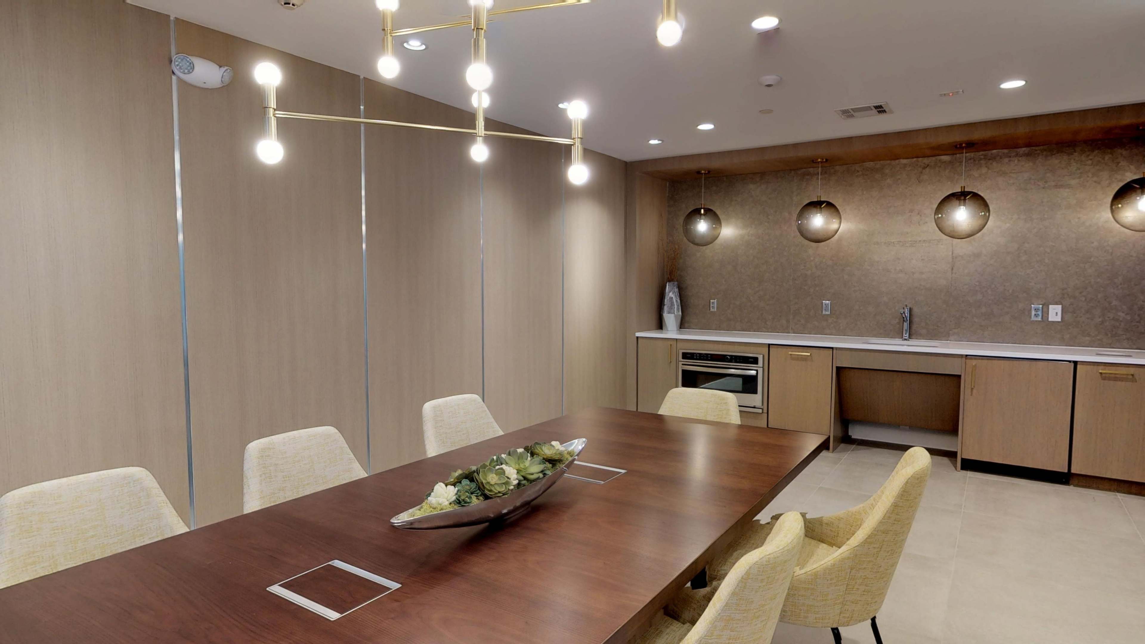 The image shows a modern conference room featuring a long wooden table surrounded by yellow upholstered chairs, with a pendant light fixture overhead and a wall-mounted sink and countertop in the background.