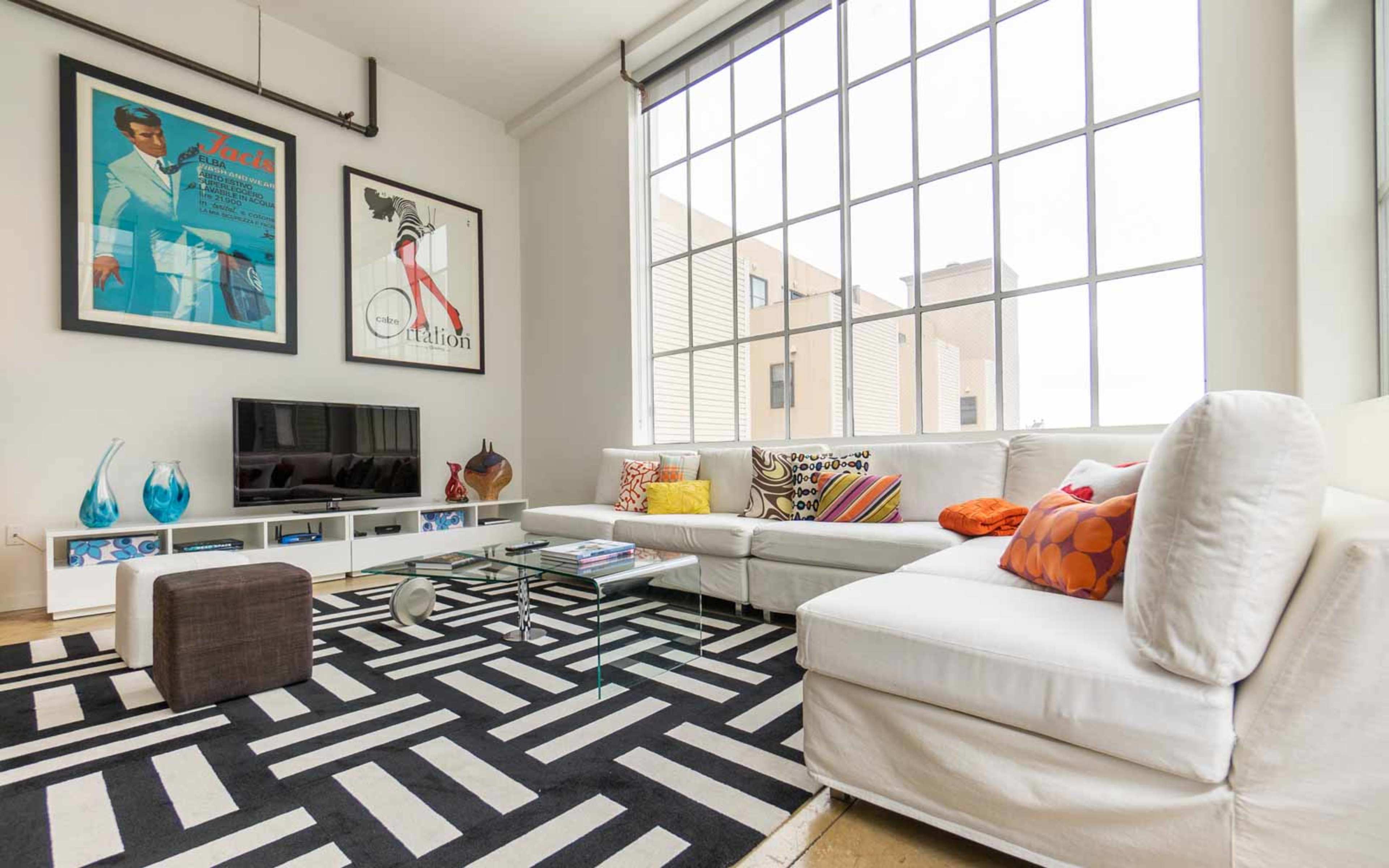 A modern living room with large windows, a white sectional sofa, a glass coffee table, and vibrant decorative pillows on a black and white patterned rug.