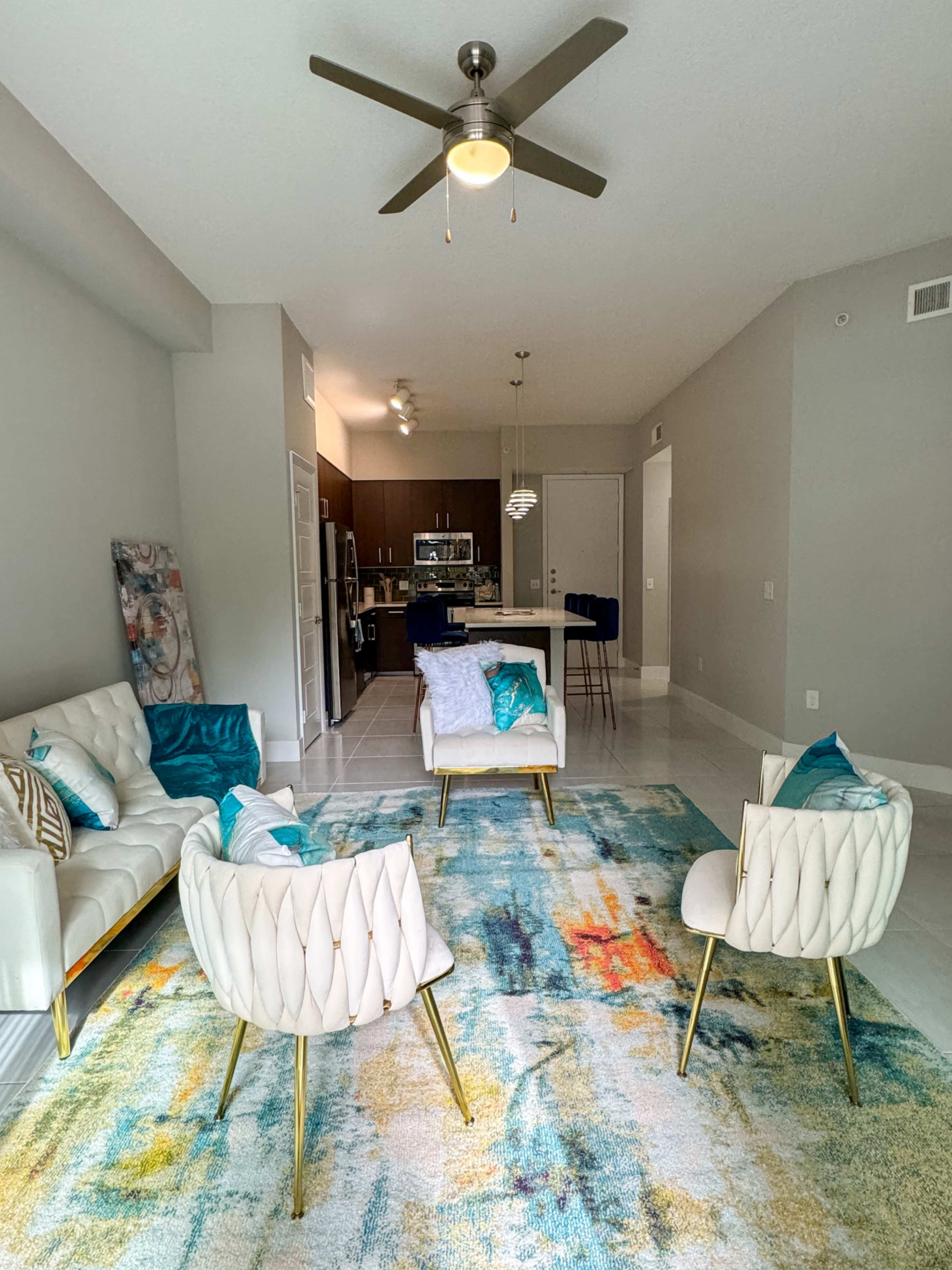 The image shows a modern living room with a white couch, two white armchairs, and a colorful rug, leading to a kitchen area in the background.