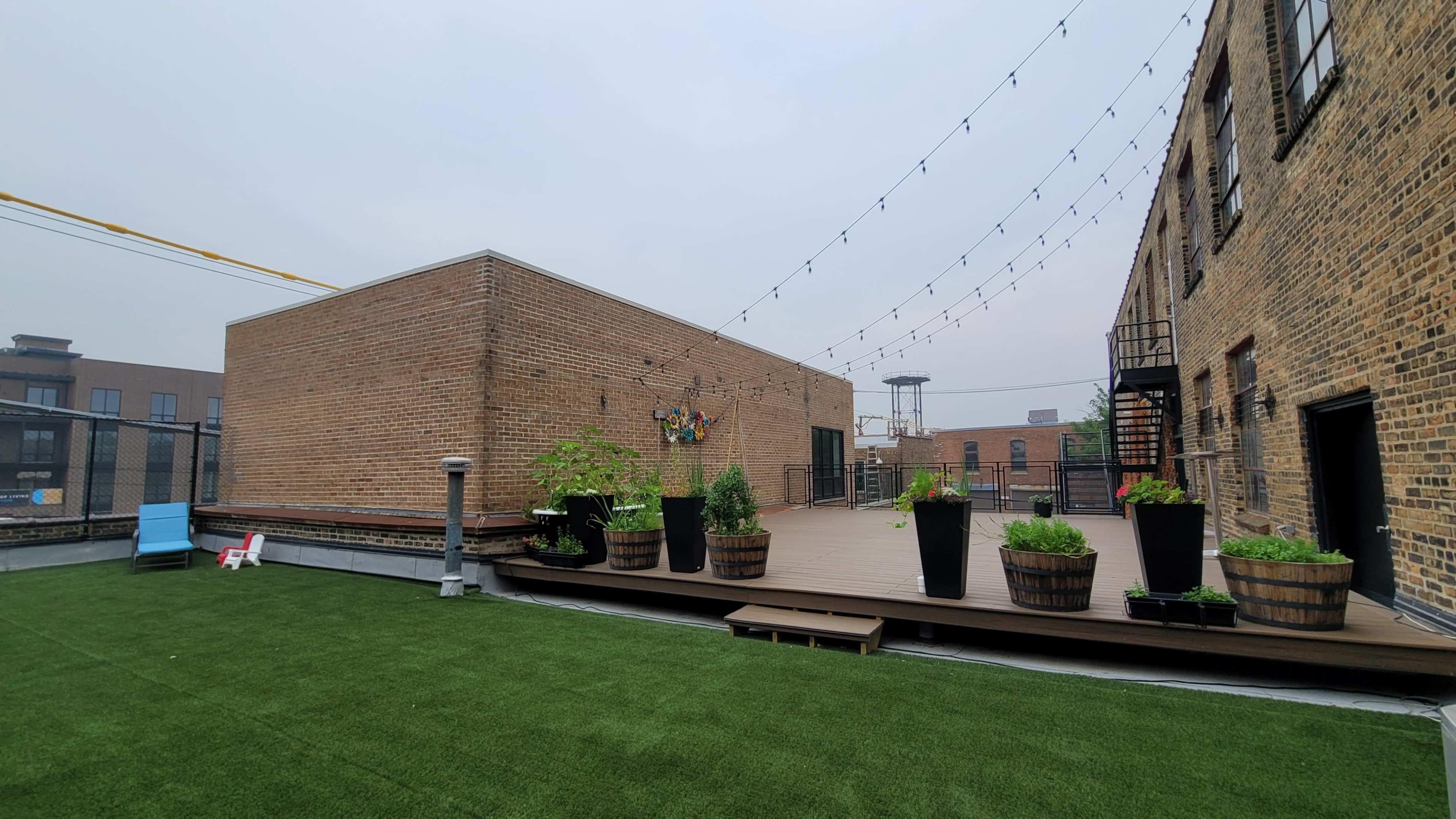 The image shows a rooftop space featuring wooden decking, large planters with greenery, and string lights overhead.