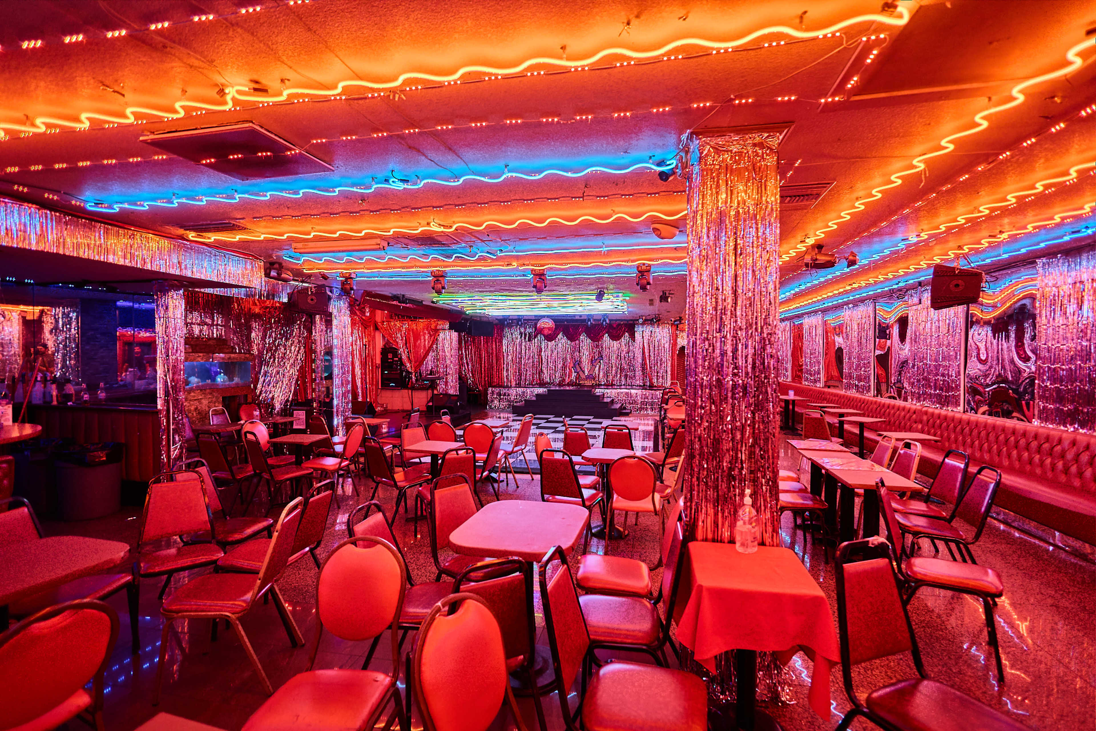 The image shows an empty, colorful night club interior with neon lights, metallic decorations, and rows of tables and chairs arranged throughout the space.