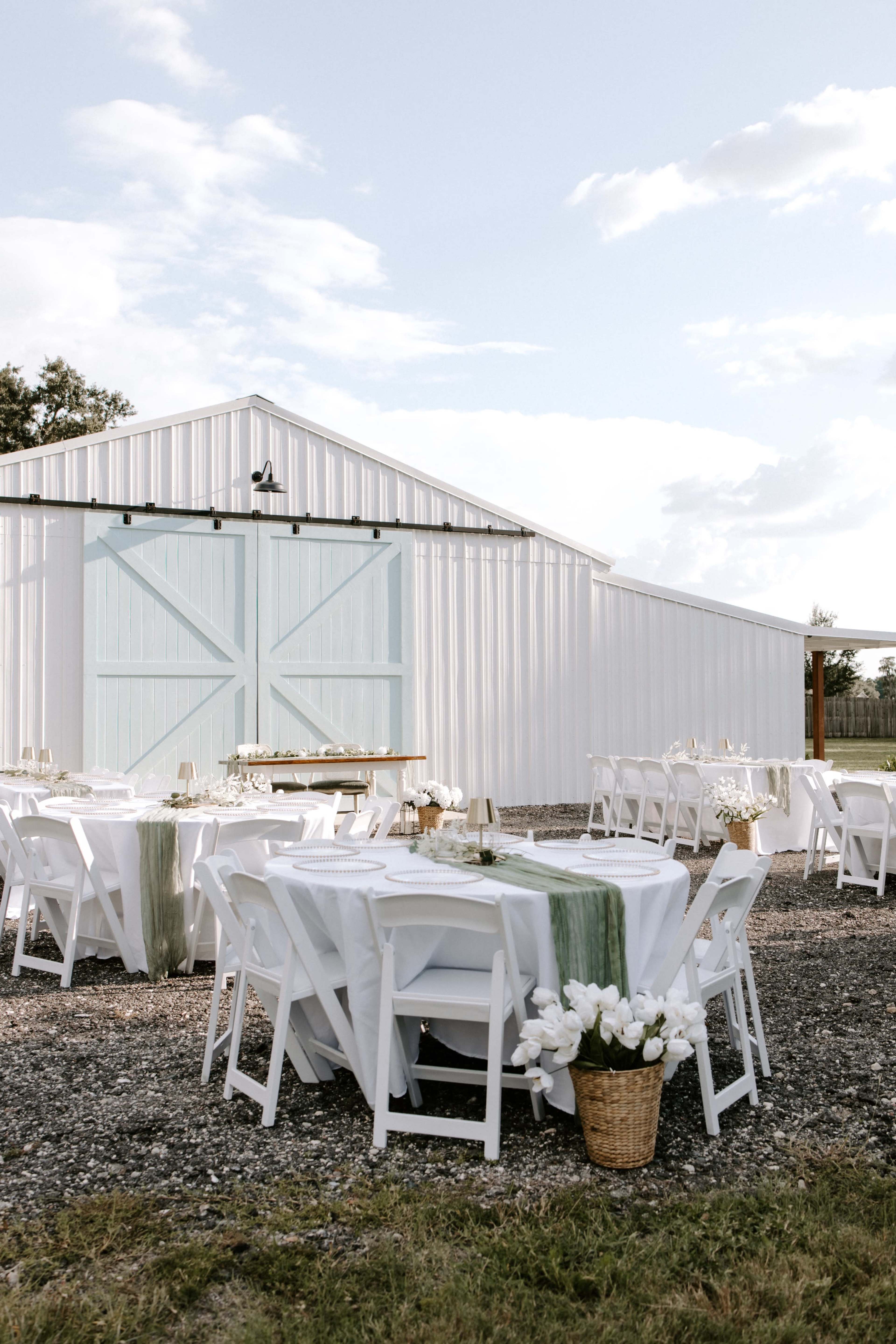 A rustic outdoor setting features white tables and chairs neatly arranged in front of a light blue barn with large sliding doors.