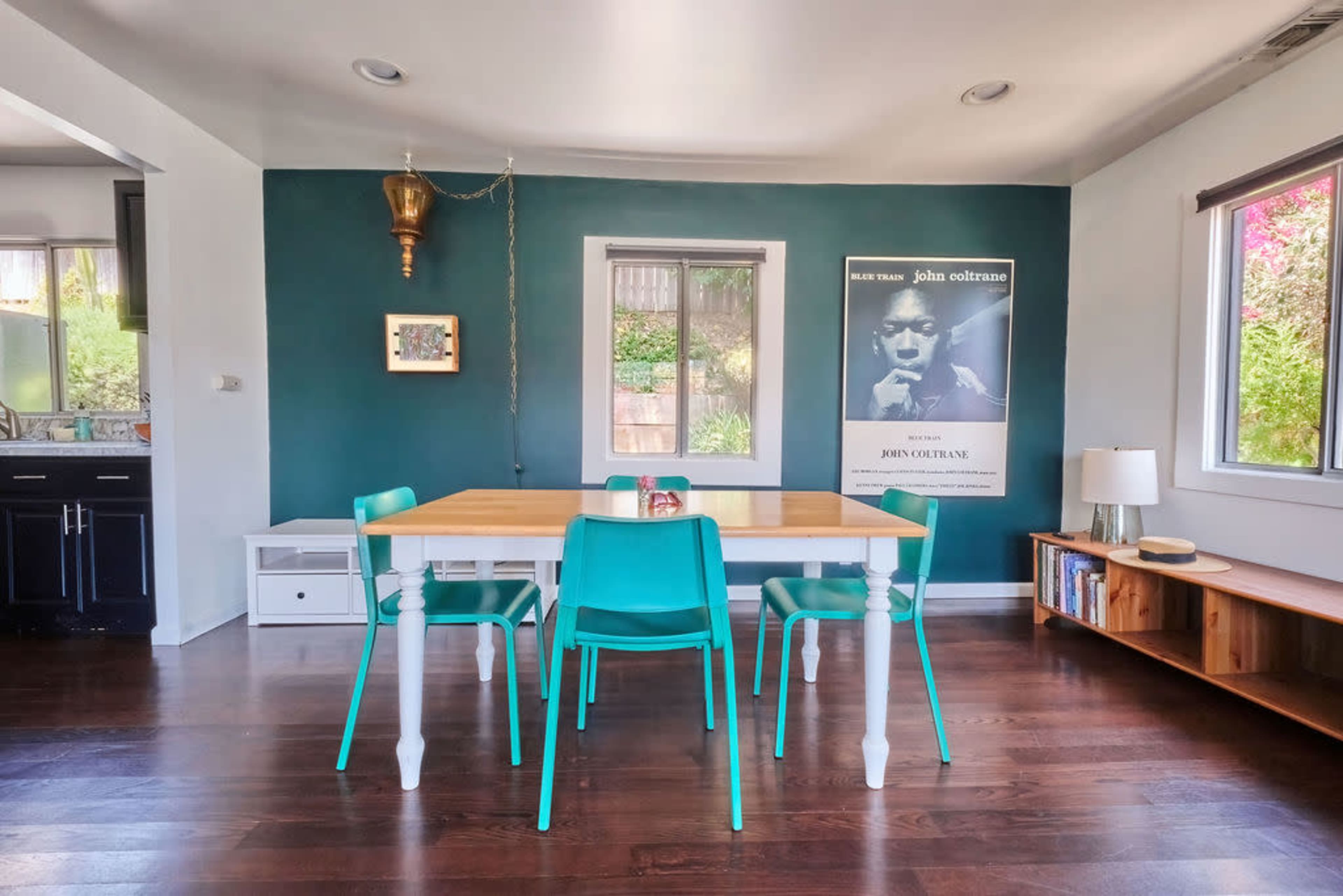 A dining area features a wooden table surrounded by turquoise chairs, with a poster of John Coltrane on the wall and a lamp on a side table.