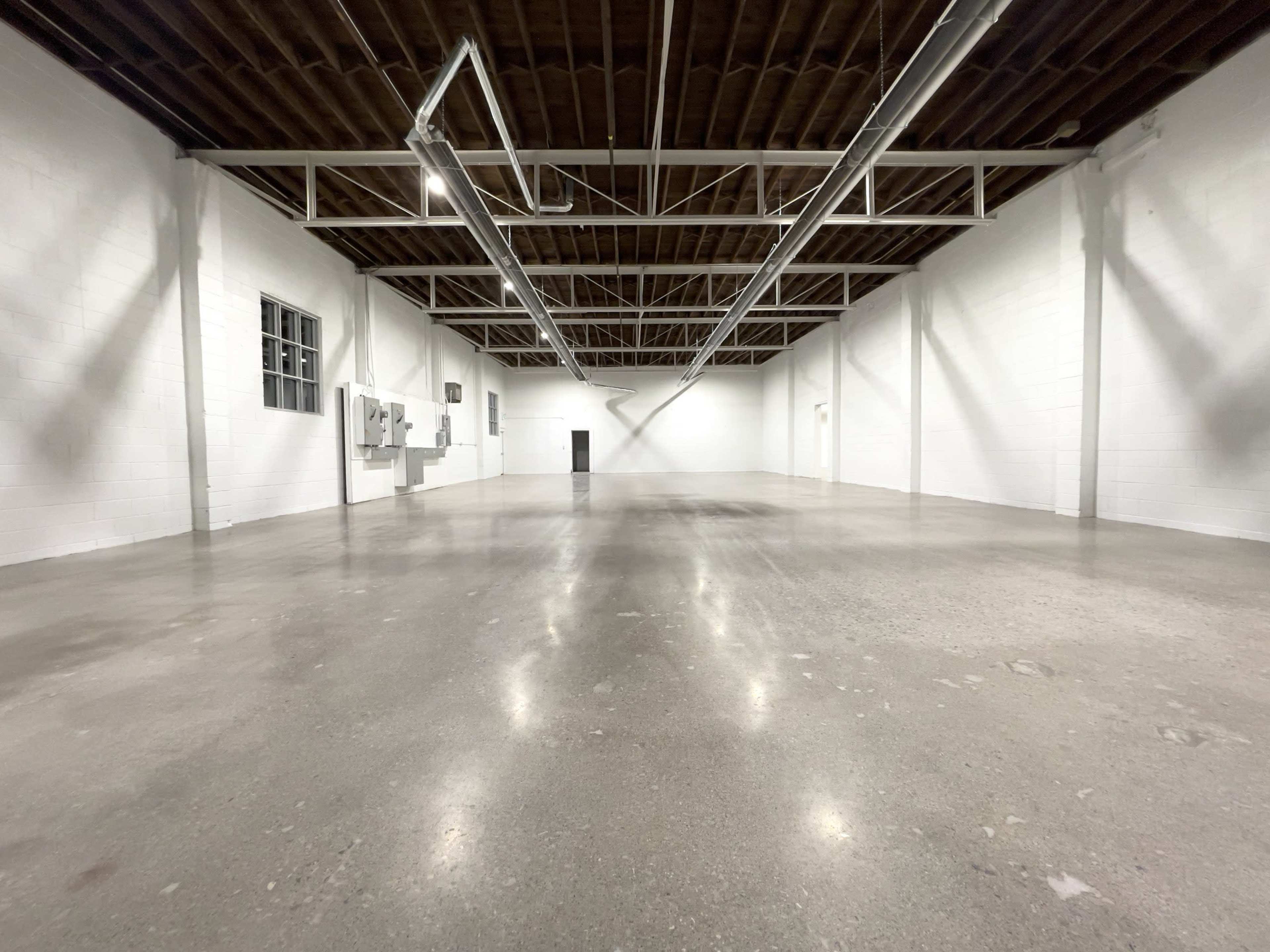 The image shows a large, empty warehouse with white walls and a polished concrete floor, featuring overhead lighting and industrial electrical panels.