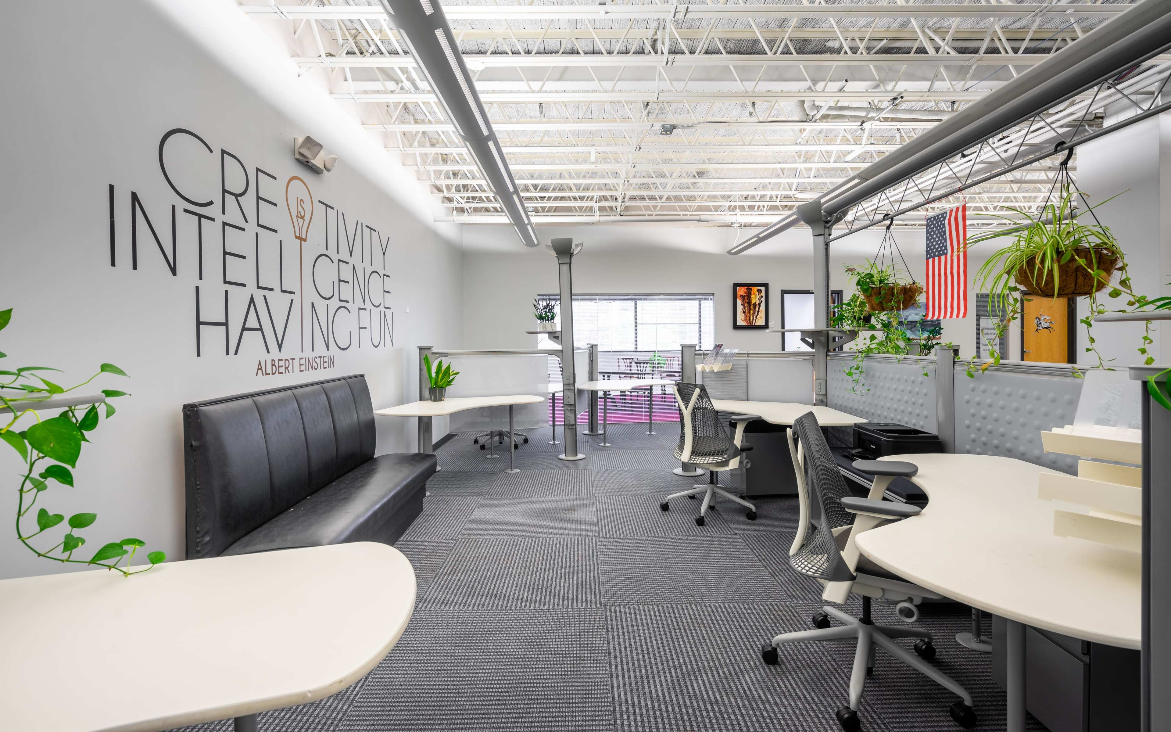 A bright office space featuring modern desks, plants, and a wall with the quote "Creativity Intelligence Having Fun" attributed to Albert Einstein.