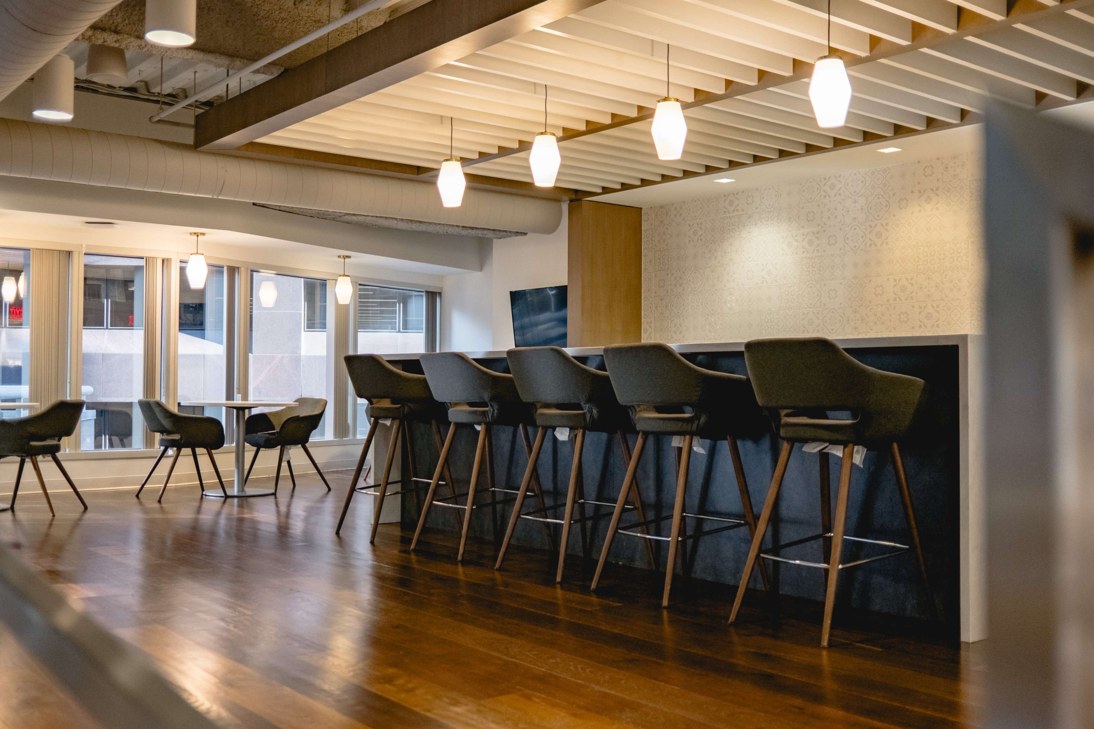 A modern interior space featuring high bar stools at a counter, with pendant lights hanging above and large windows providing natural light.