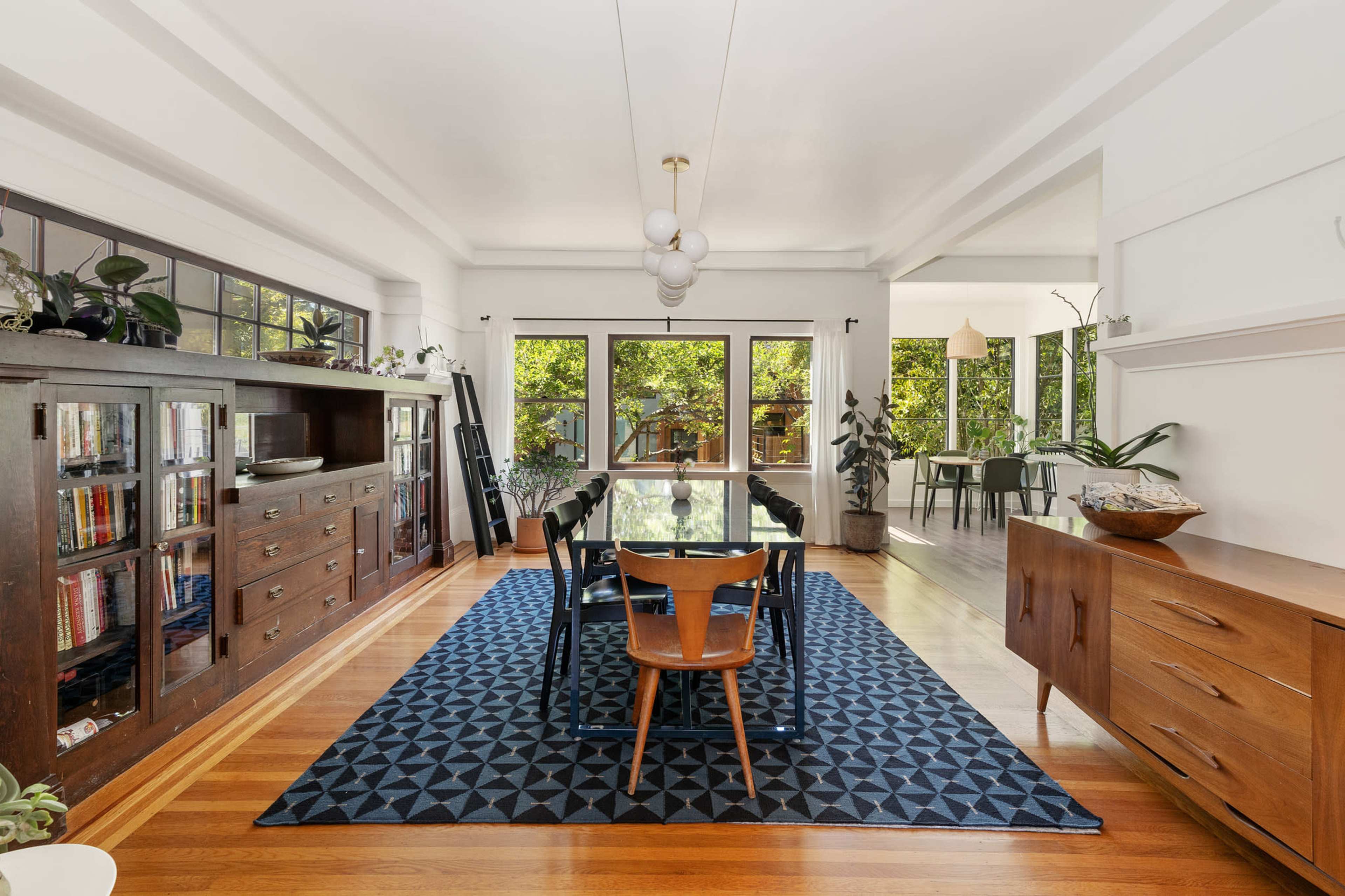 A dining area features a rectangular table surrounded by chairs, with large windows revealing a view of greenery outside.