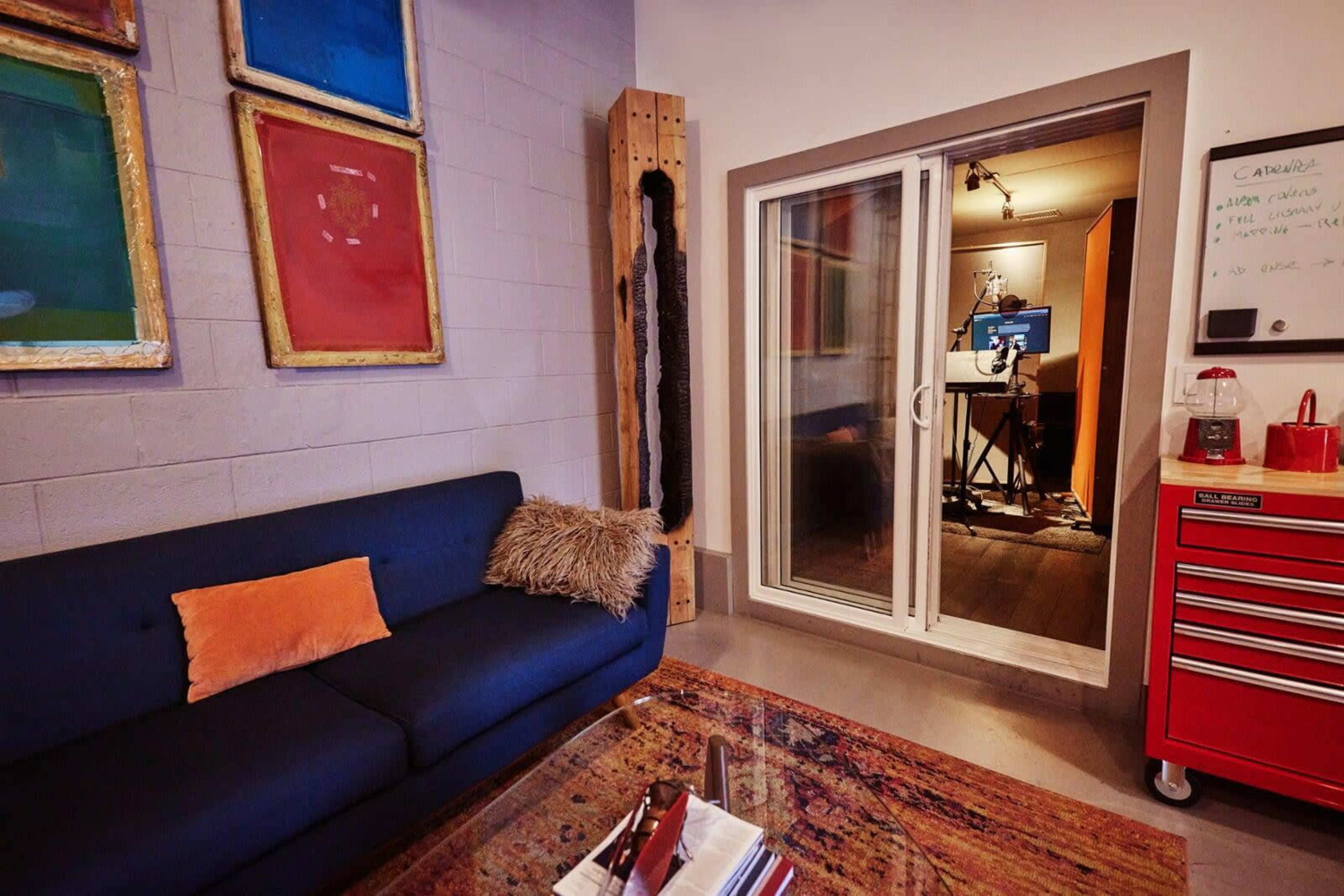 A modern room features a dark sofa with a decorative pillow, colorful framed art on the wall, and a glass door leading to another space with a recording setup visible.