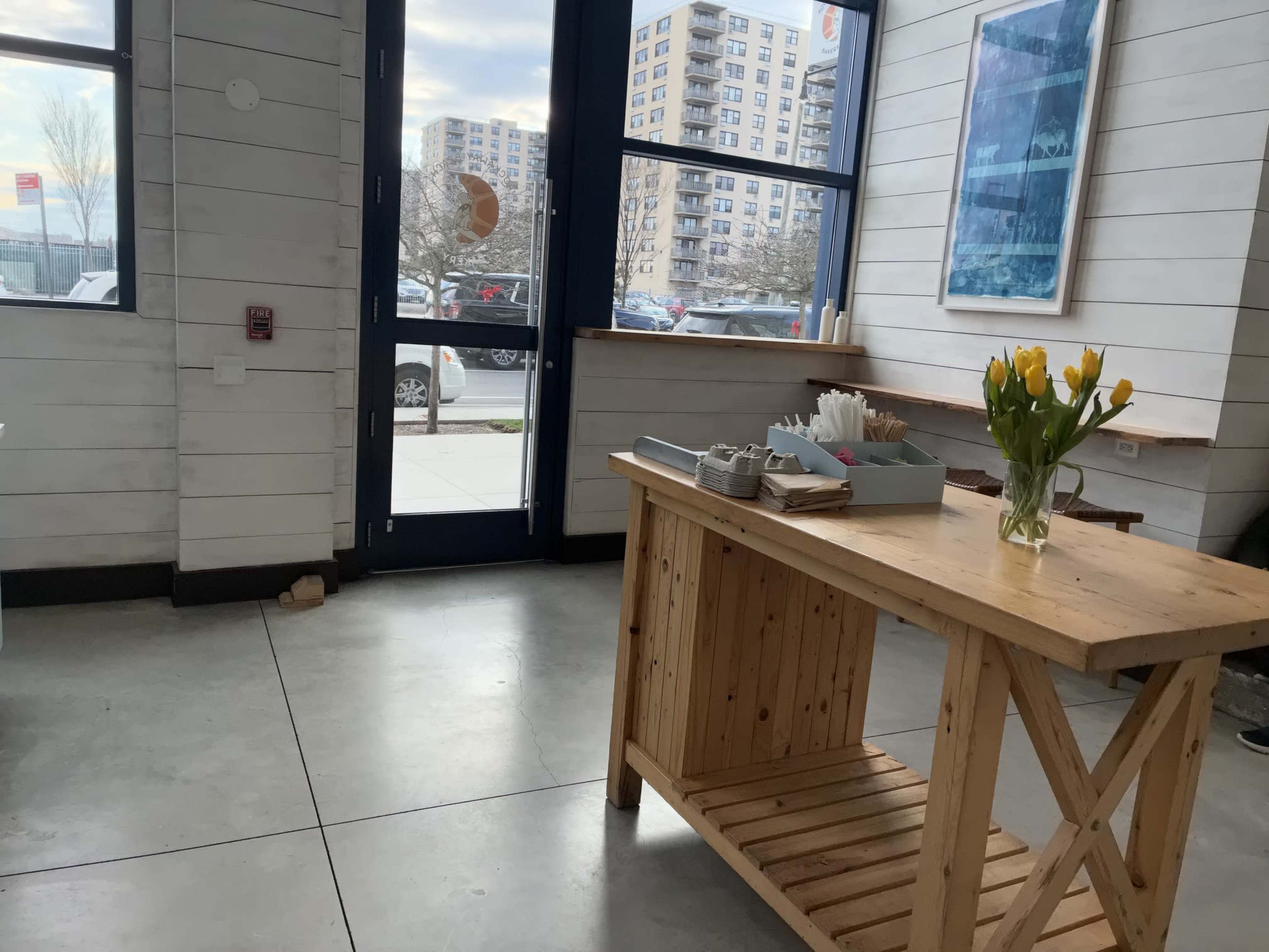 The image shows a wooden table with a vase of yellow tulips, situated near a large window in a light-colored room.