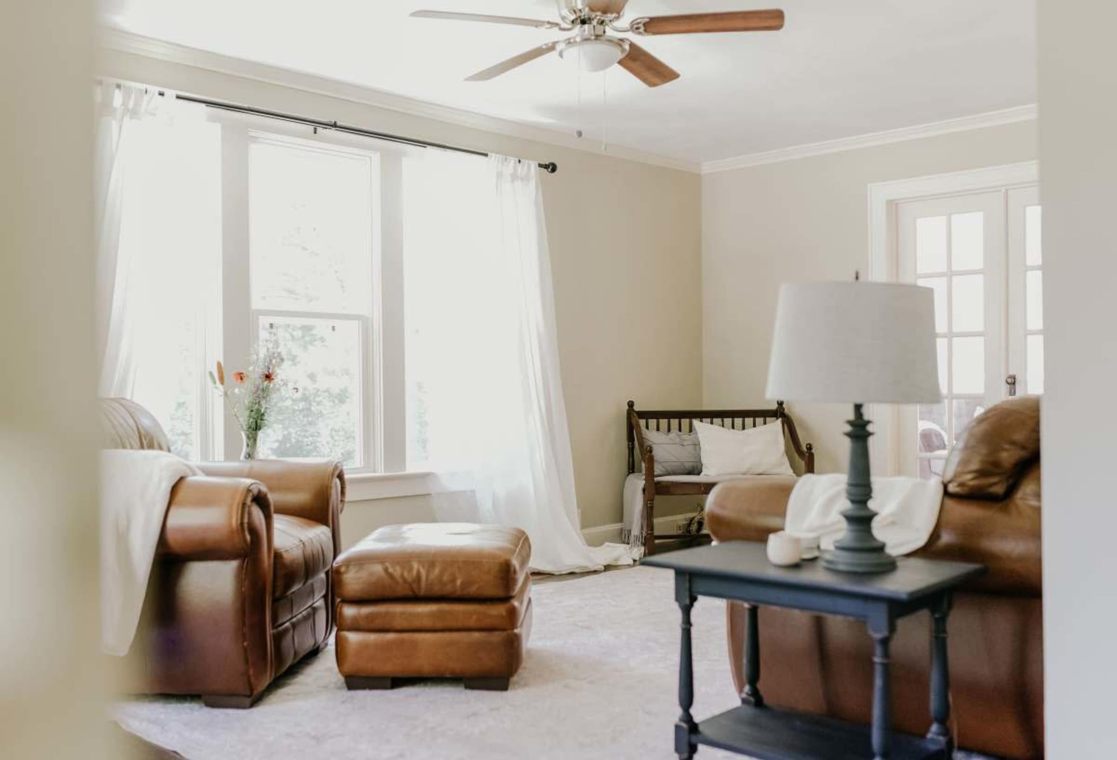 A cozy living room features brown leather furniture, a ceiling fan, large windows with sheer curtains, and a small table with a lamp.