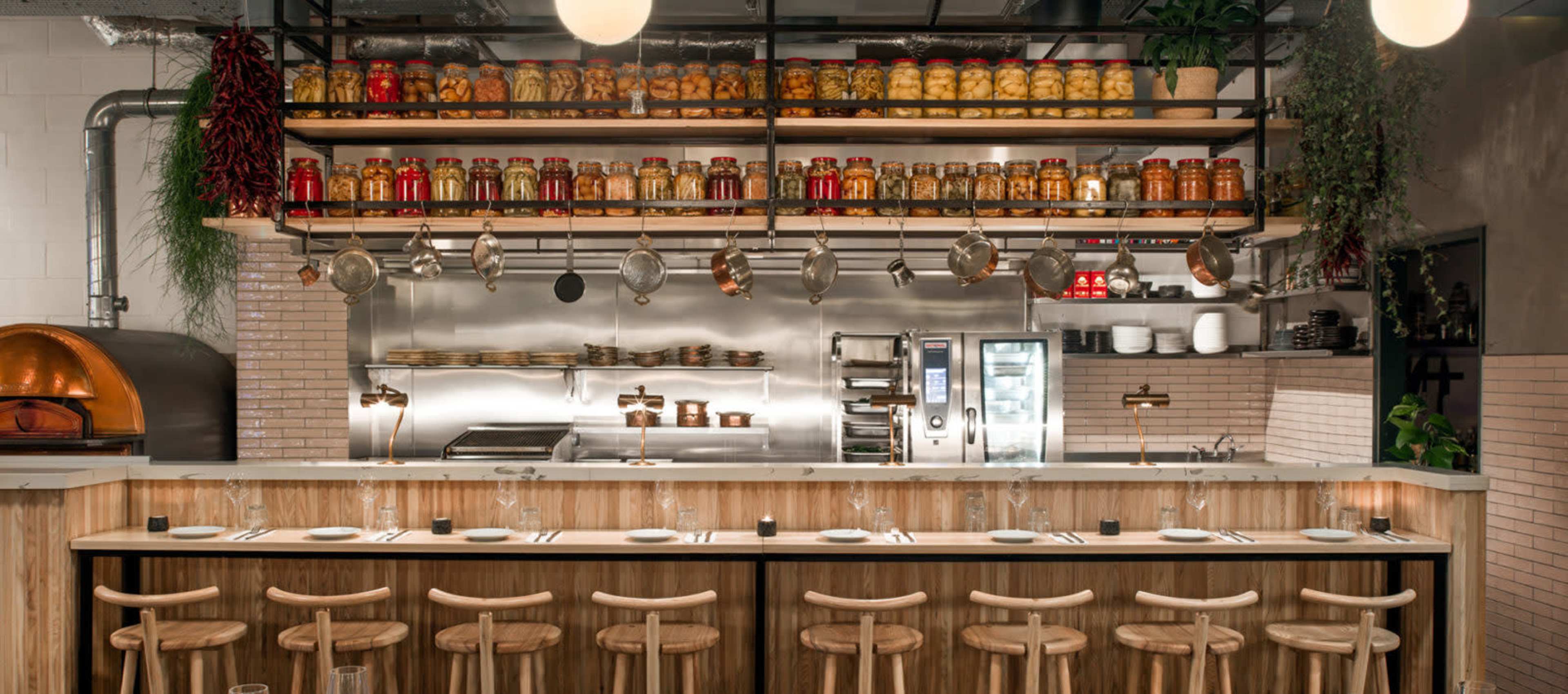 The image shows a modern restaurant kitchen with a wooden bar, shelves lined with jars of preserved ingredients, and a stainless steel work area equipped with cooking appliances.