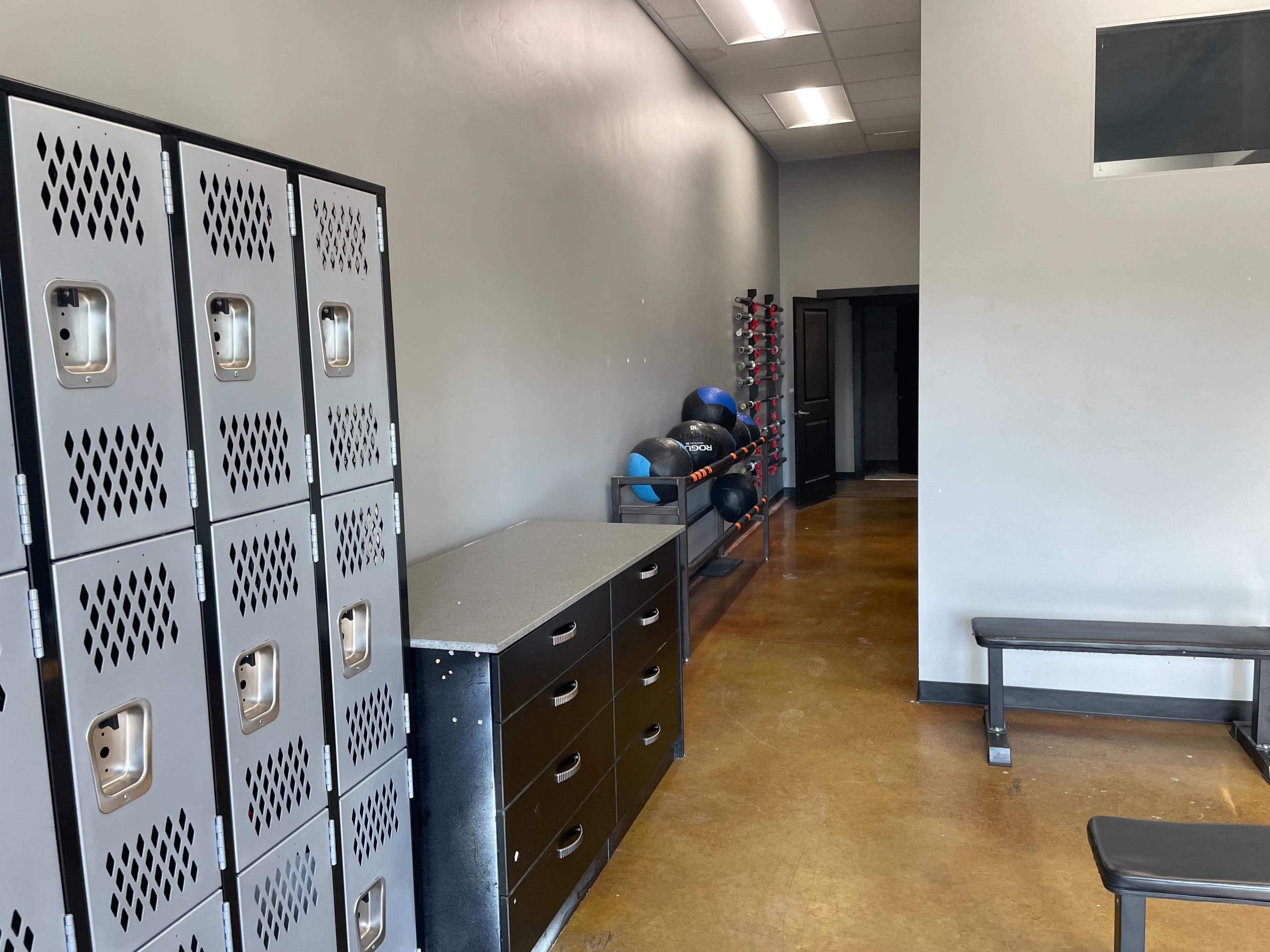 A gym locker room featuring metallic lockers, a black storage cabinet, exercise balls on a rack, and a weight bench along the wall.