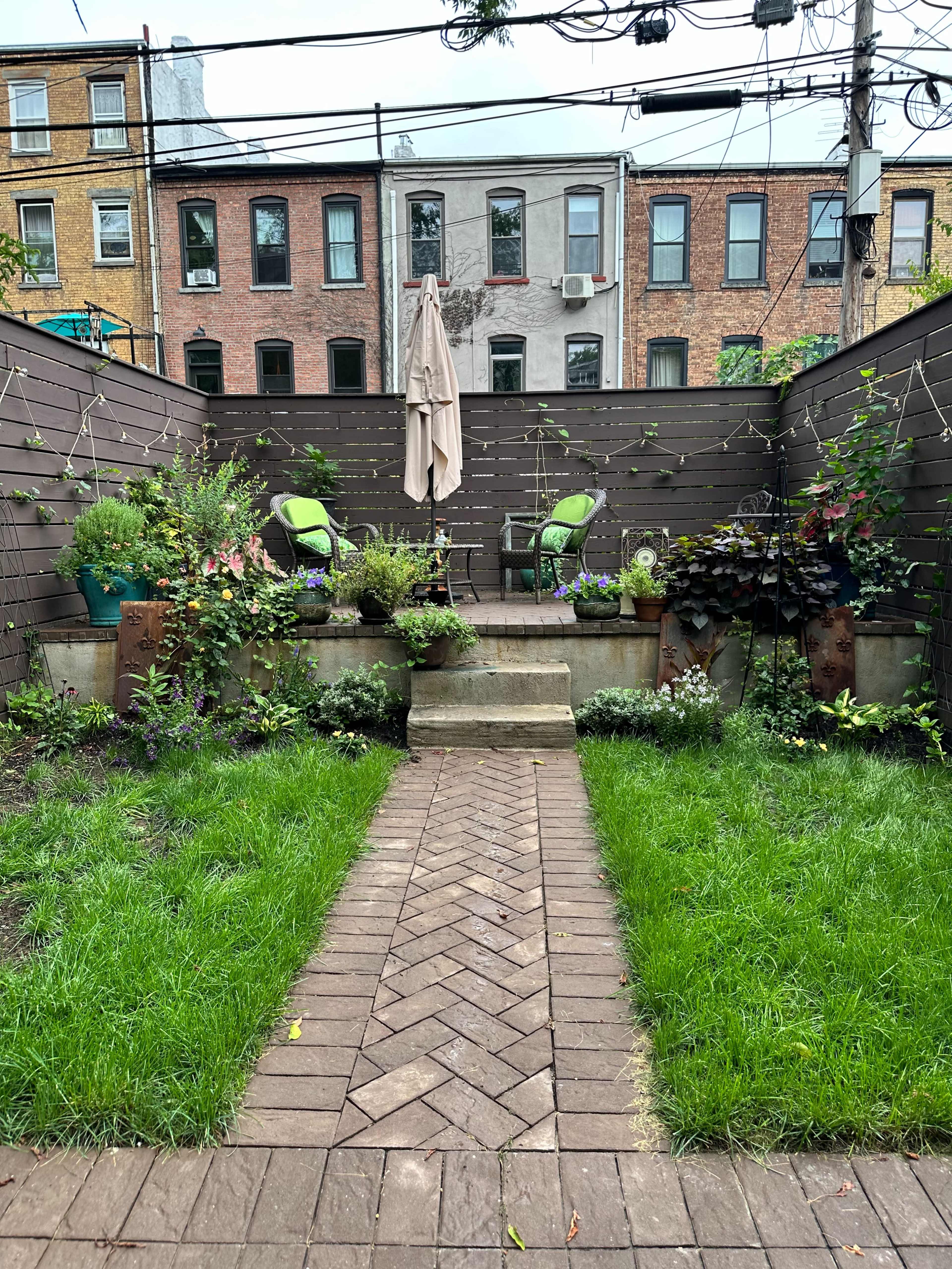The image shows a small backyard garden featuring two green chairs, a large umbrella, a stone pathway, and various plants and flowers.