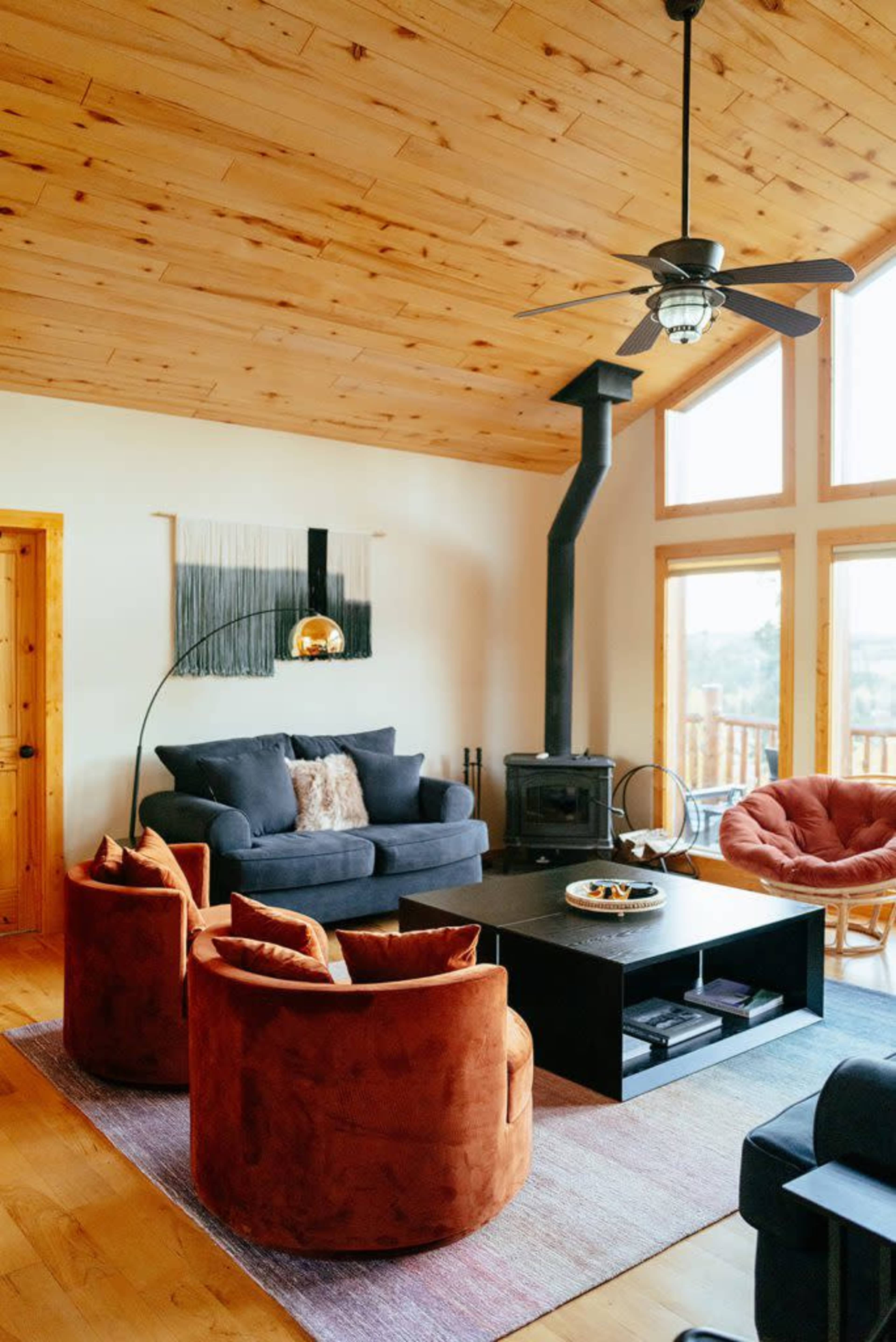The image shows a modern living room with a wooden ceiling, a black wood stove, a black coffee table, and orange accent chairs arranged around it, with large windows letting in natural light.