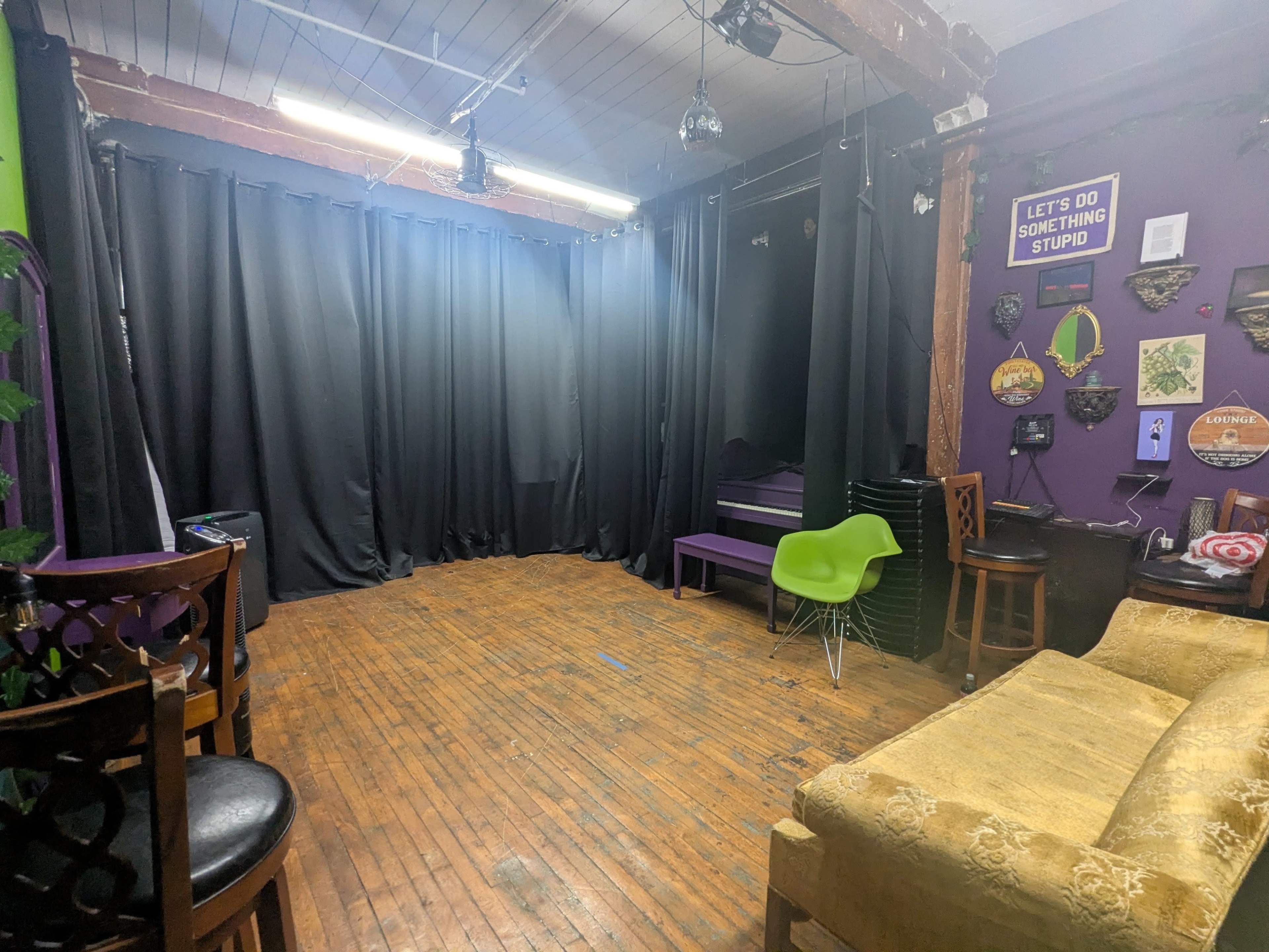 The image shows a dimly lit room with wooden floors, black curtains along one wall, and various pieces of furniture, including green chairs and a couch.