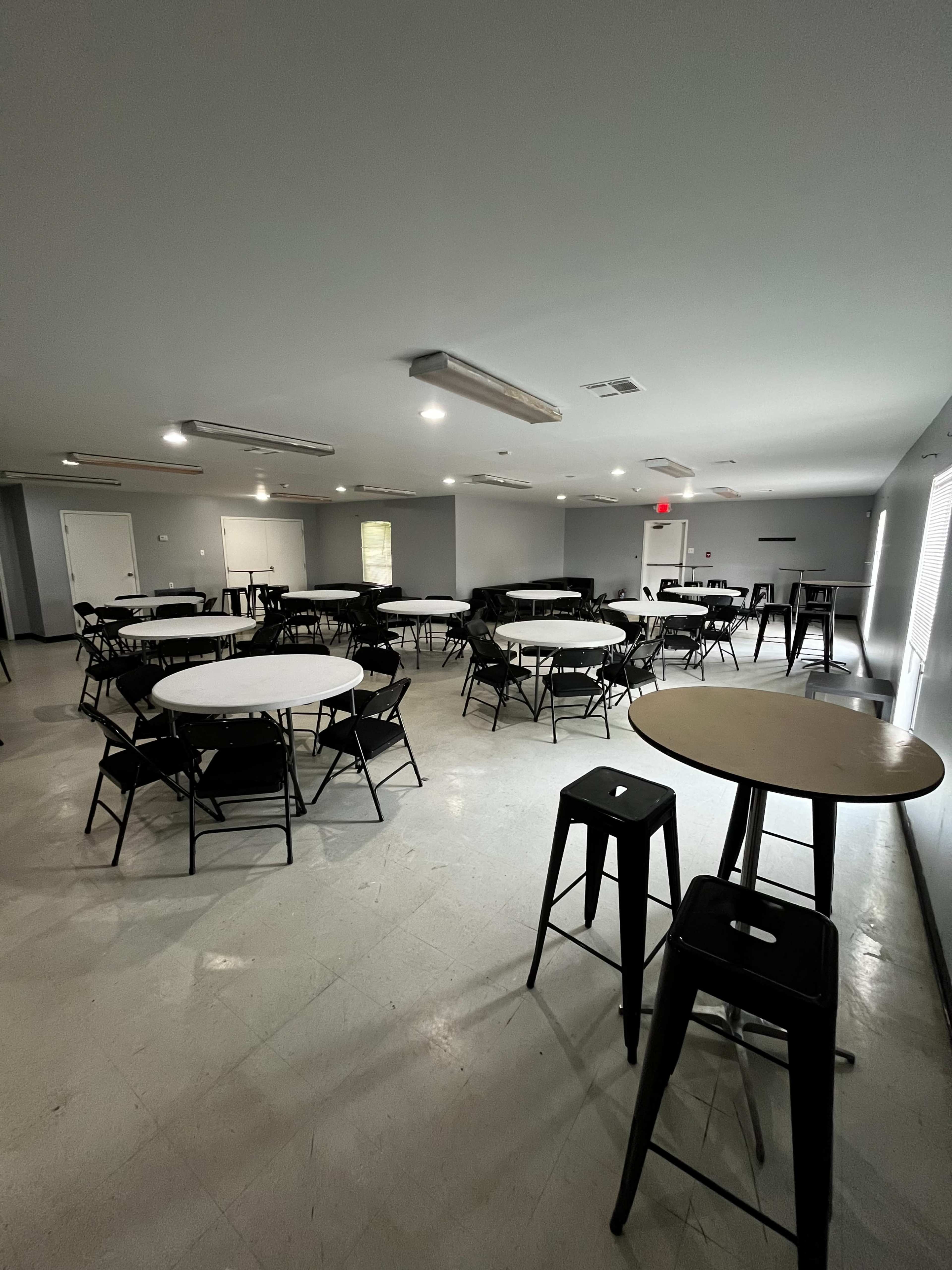 The image shows a spacious room with several round tables and black chairs arranged throughout, illuminated by overhead lights.