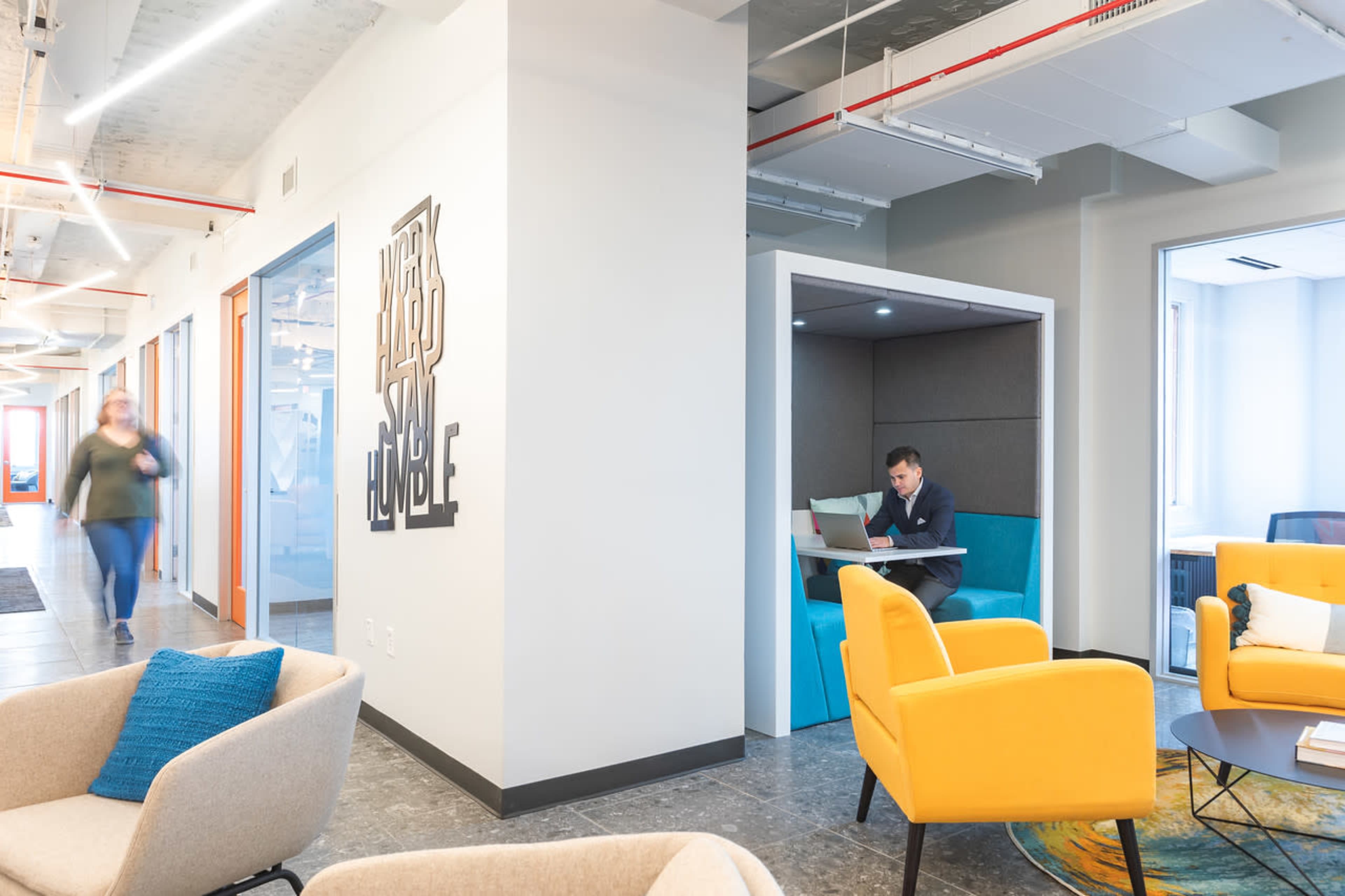 A modern office space features a seating area with bright chairs, a private booth occupied by a person working on a laptop, and a person walking in the background.