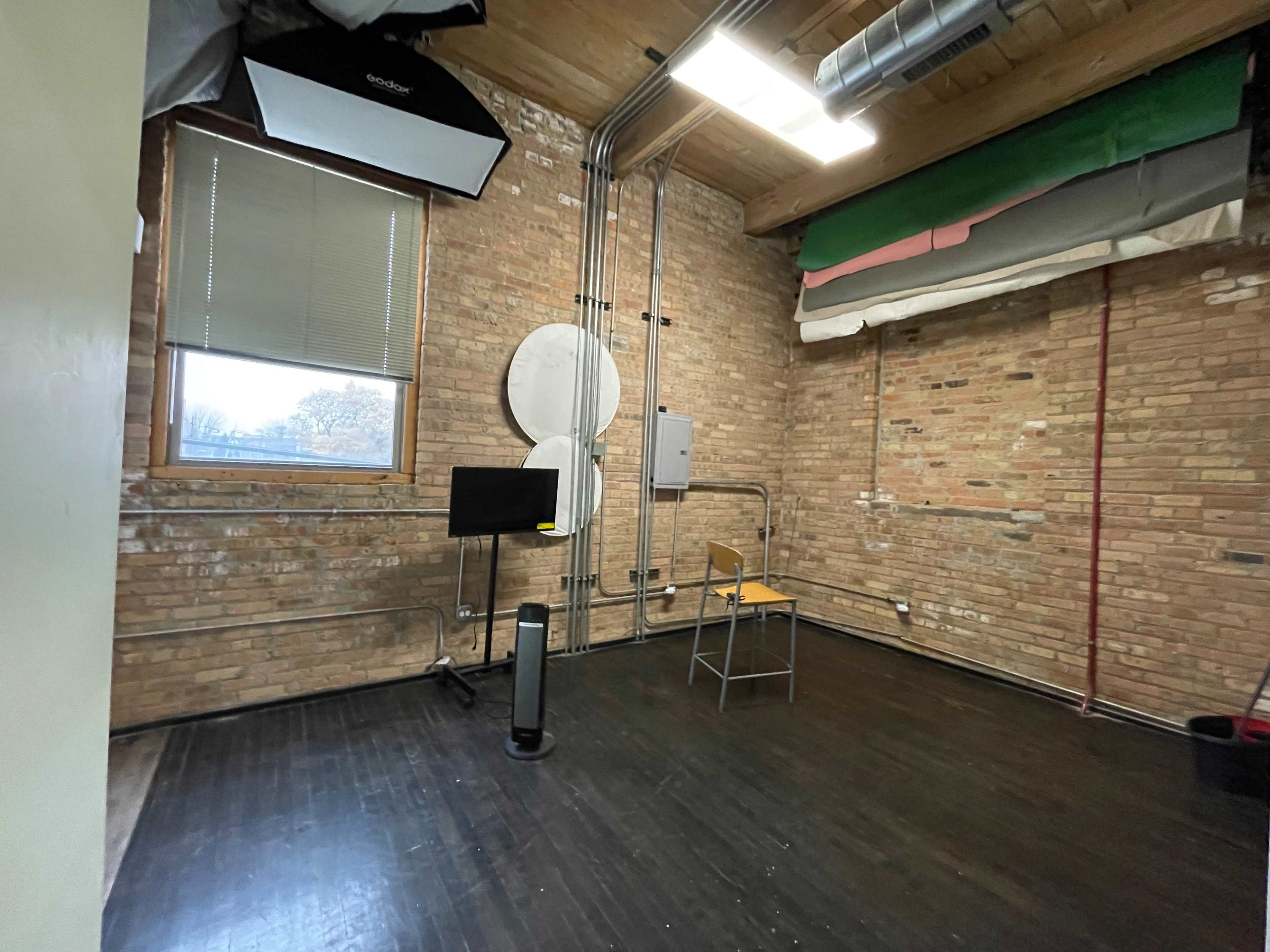 The image shows an empty room with exposed brick walls, wooden flooring, a chair, a monitor on a stand, and visible electrical piping.