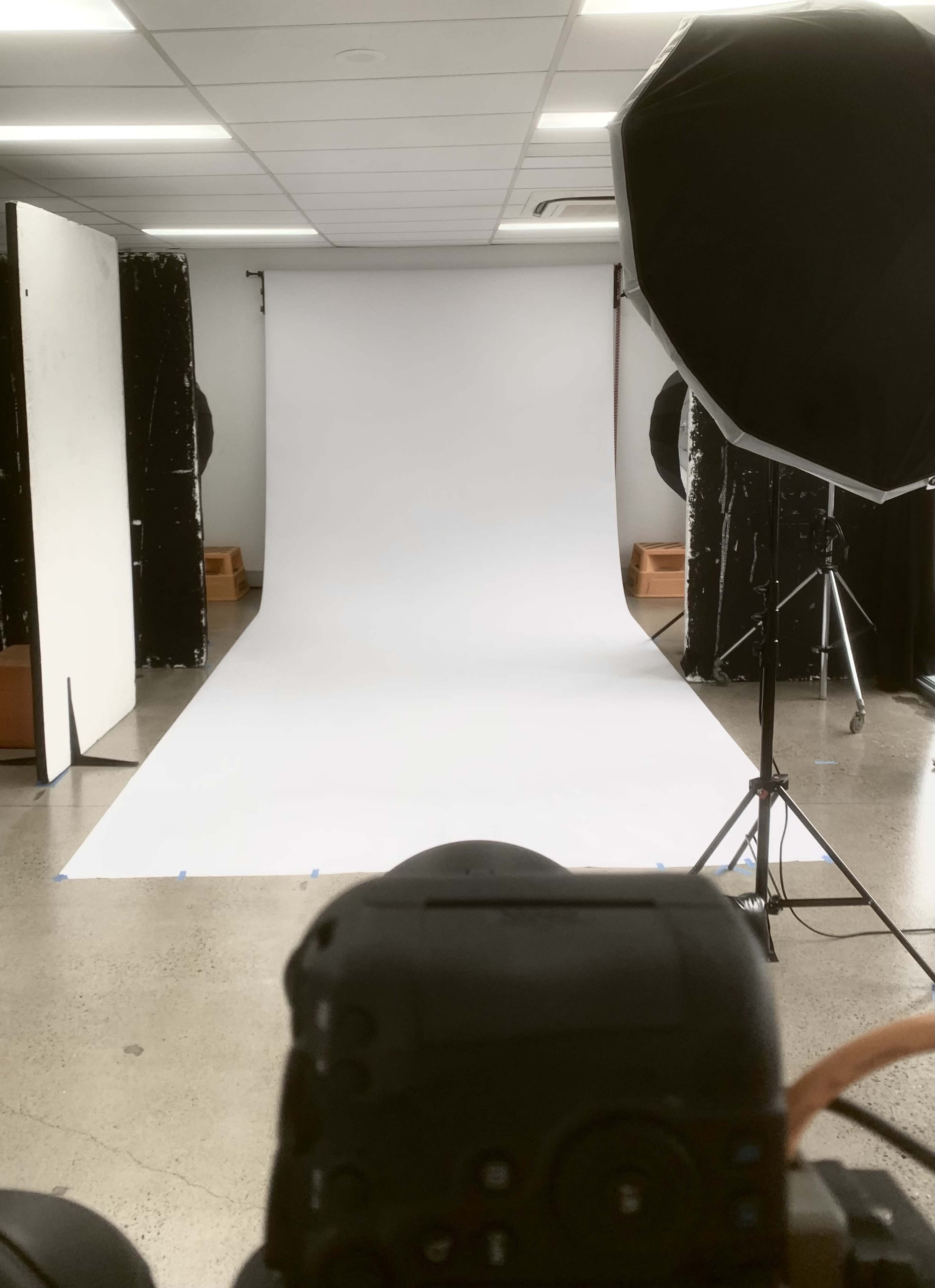 A photography studio with a white backdrop, a camera positioned in the foreground, and a large softbox light setup.