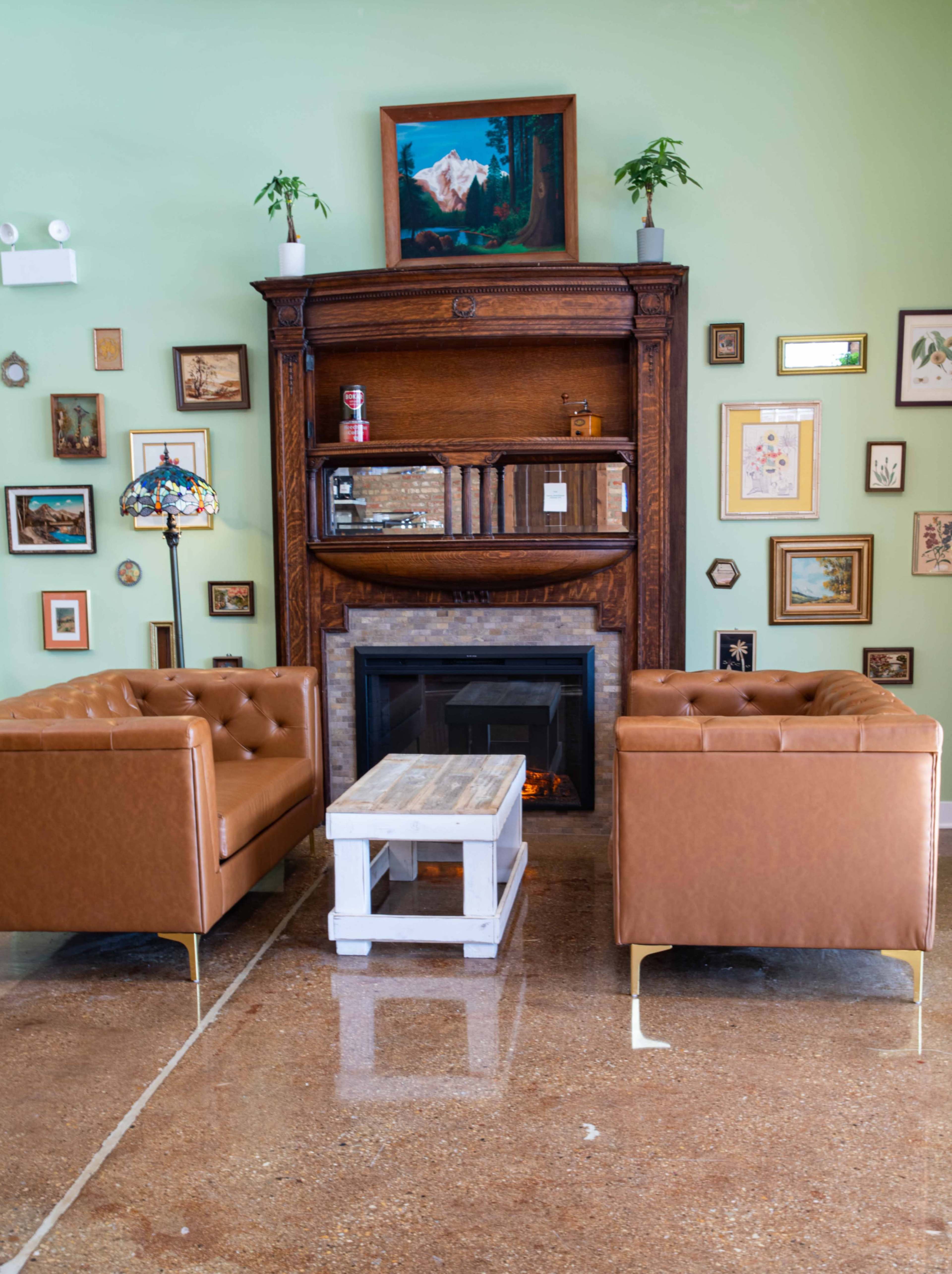 A cozy lounge area features two brown leather chairs facing a vintage wooden fireplace, surrounded by a collection of framed artwork on the green walls.