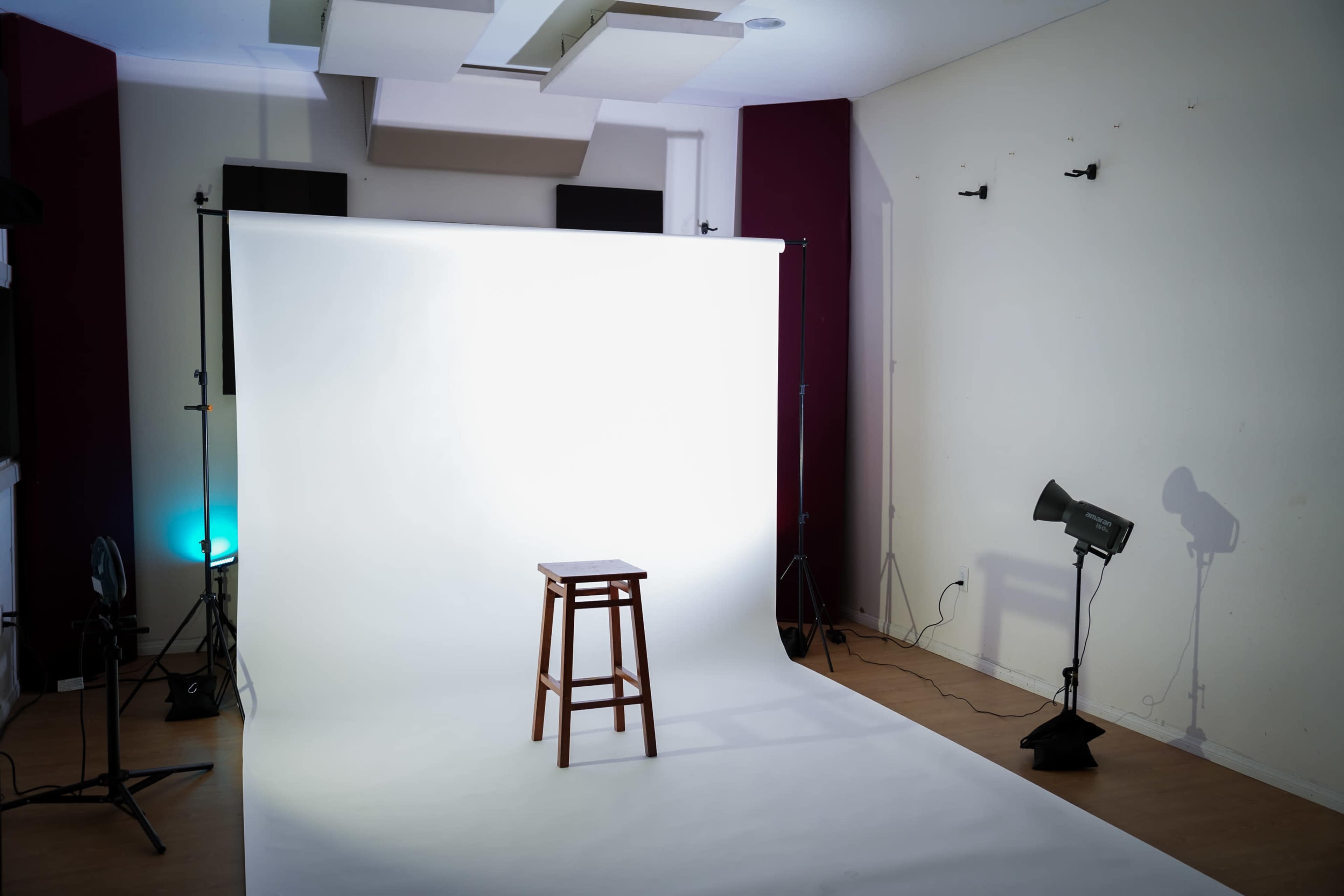 The image shows a photography studio setup with a plain white backdrop, a wooden stool in the center, and several lighting fixtures around the space.