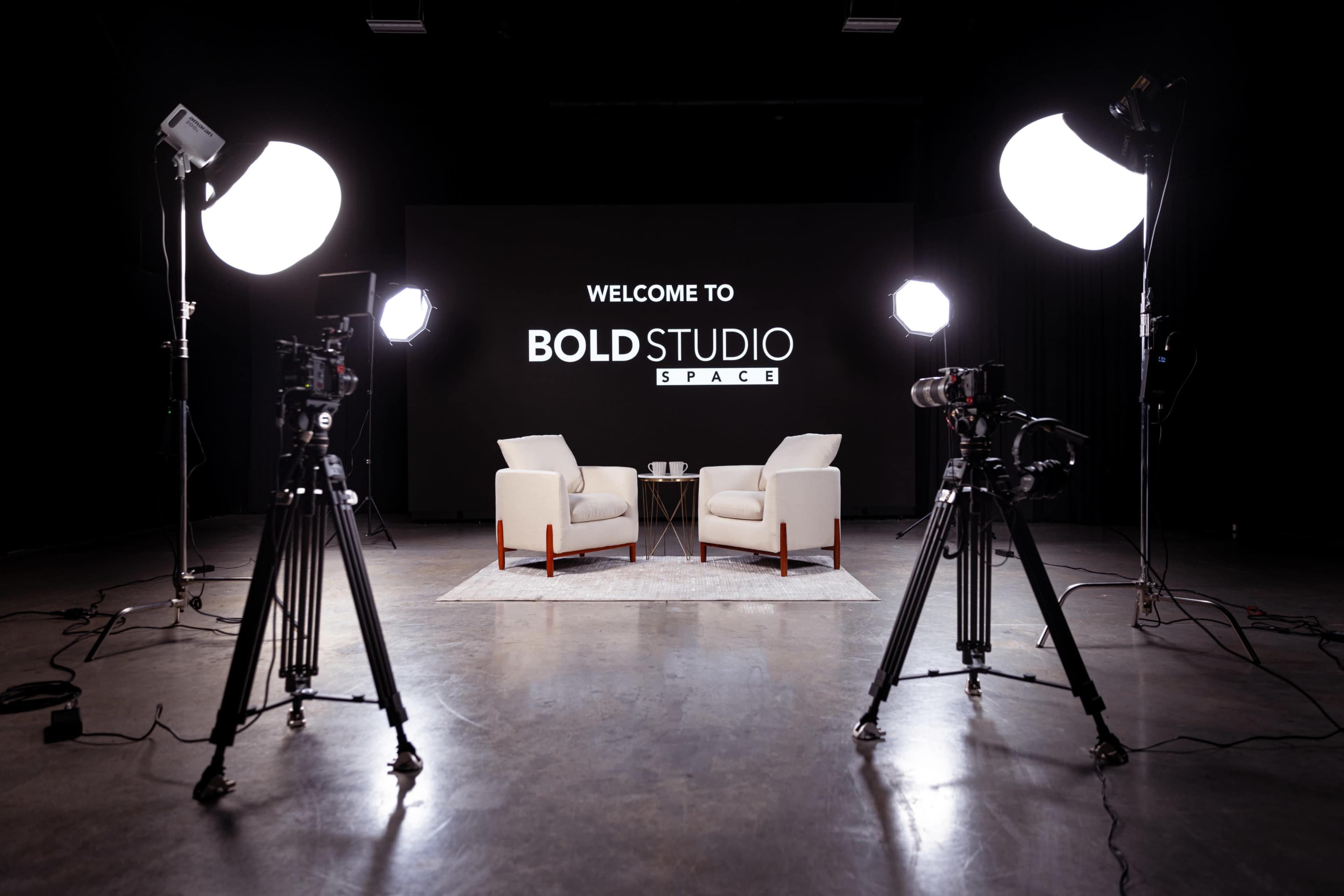 A well-lit studio space featuring two white chairs facing each other, with a small table in between and professional cameras on tripods set up nearby.