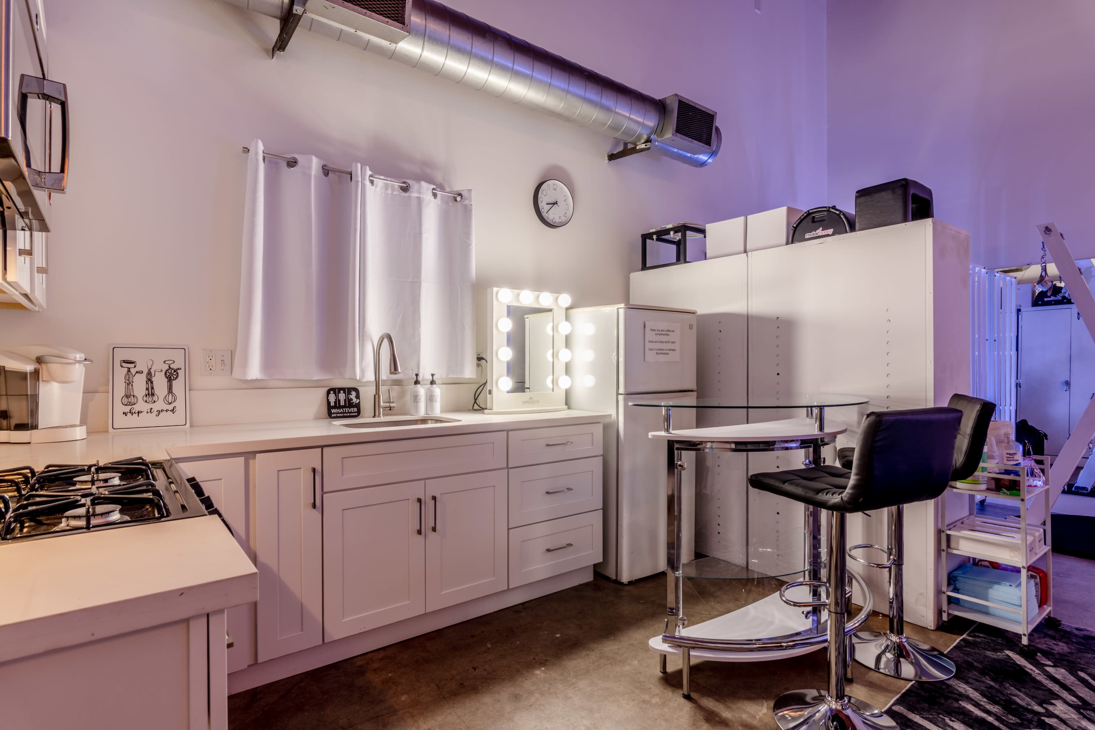 The image shows a modern kitchenette with white cabinetry, a sink, and a round bar table with a chair in an industrial-style setting.