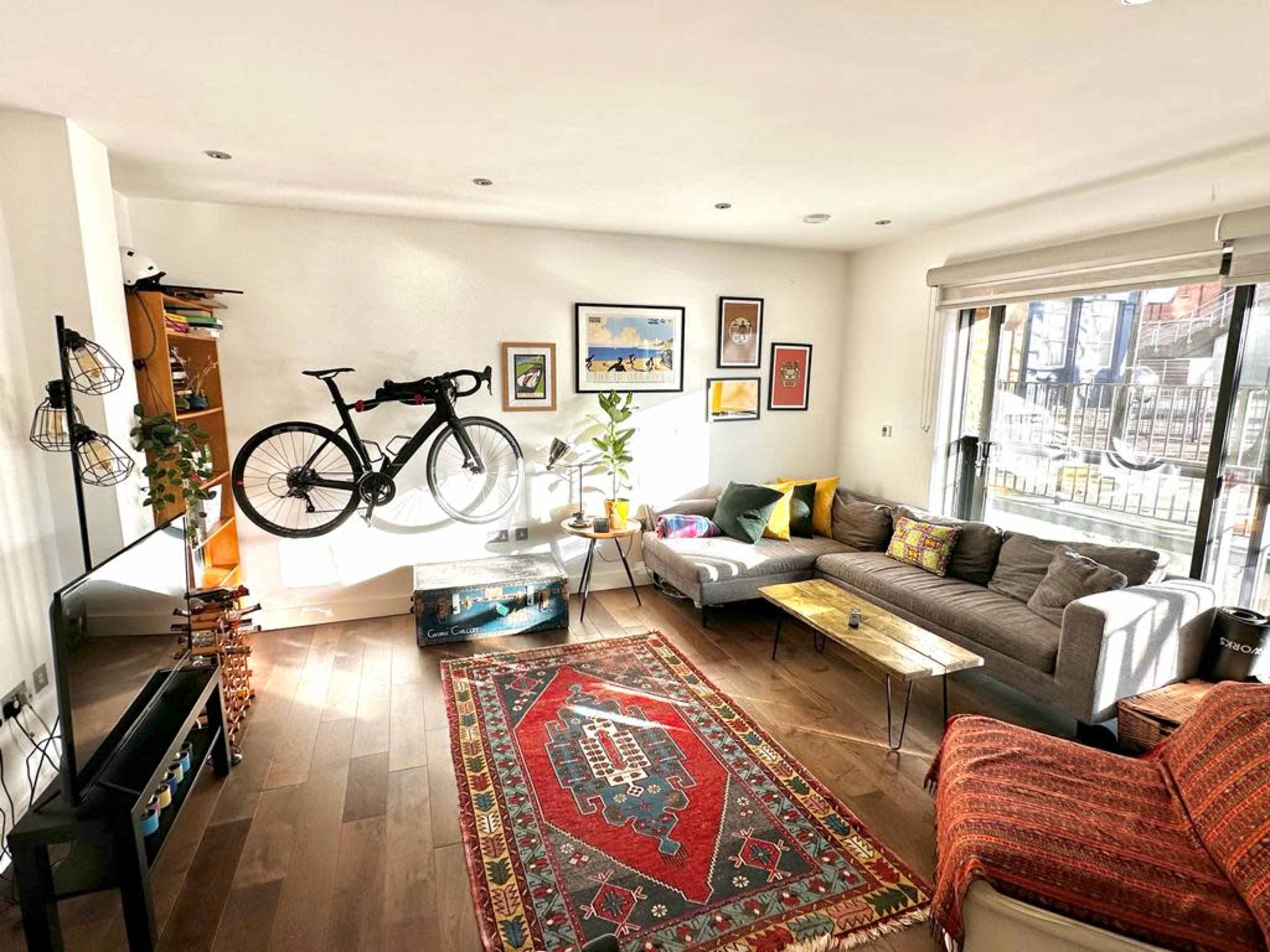 A modern living room featuring a gray sectional sofa, a bicycle mounted on the wall, a rug on the wooden floor, and large windows allowing natural light to fill the space.