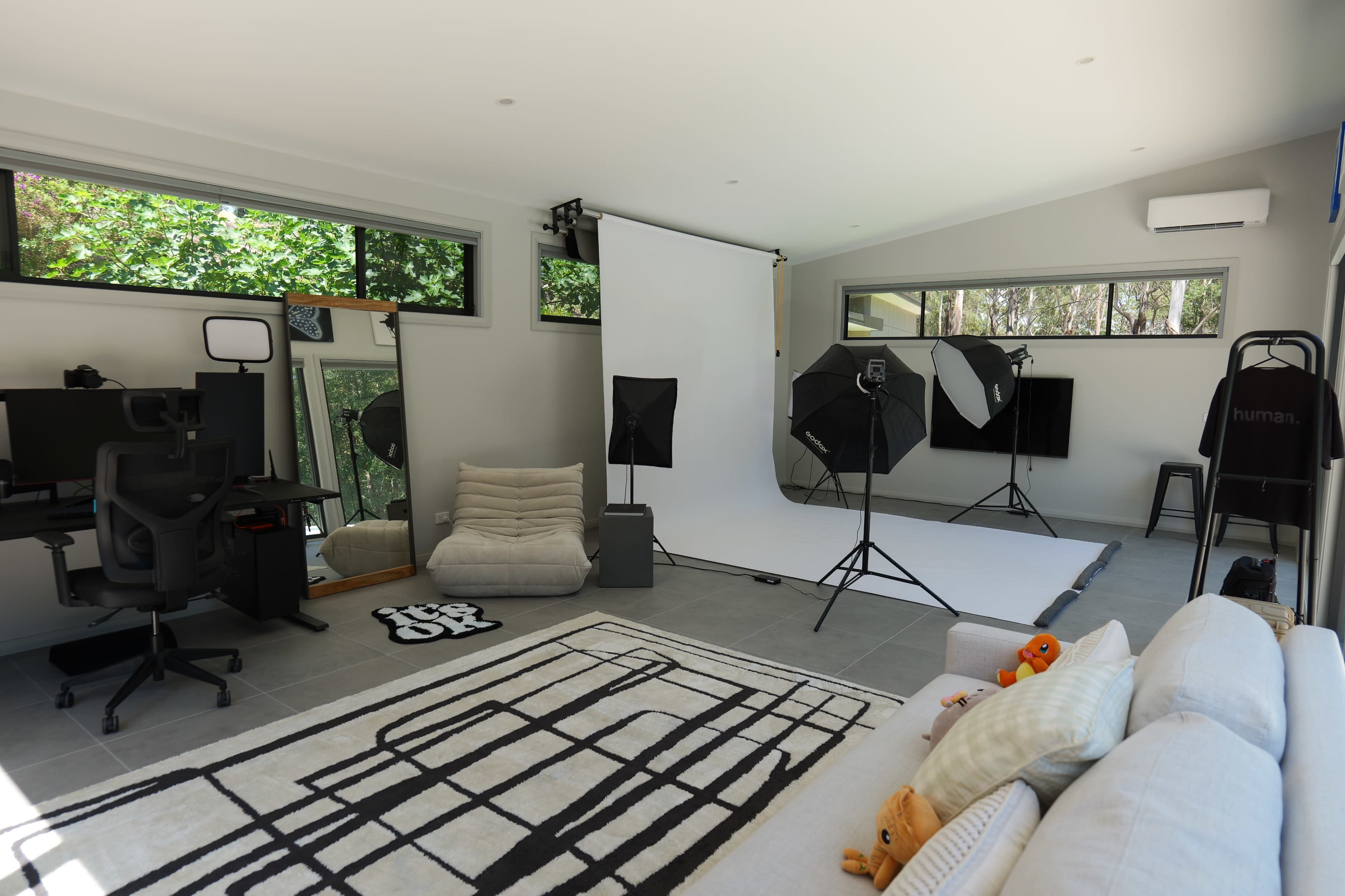 The image shows a spacious studio with a white backdrop, soft lighting equipment, and a cozy seating area, along with a modern workspace and a patterned rug.