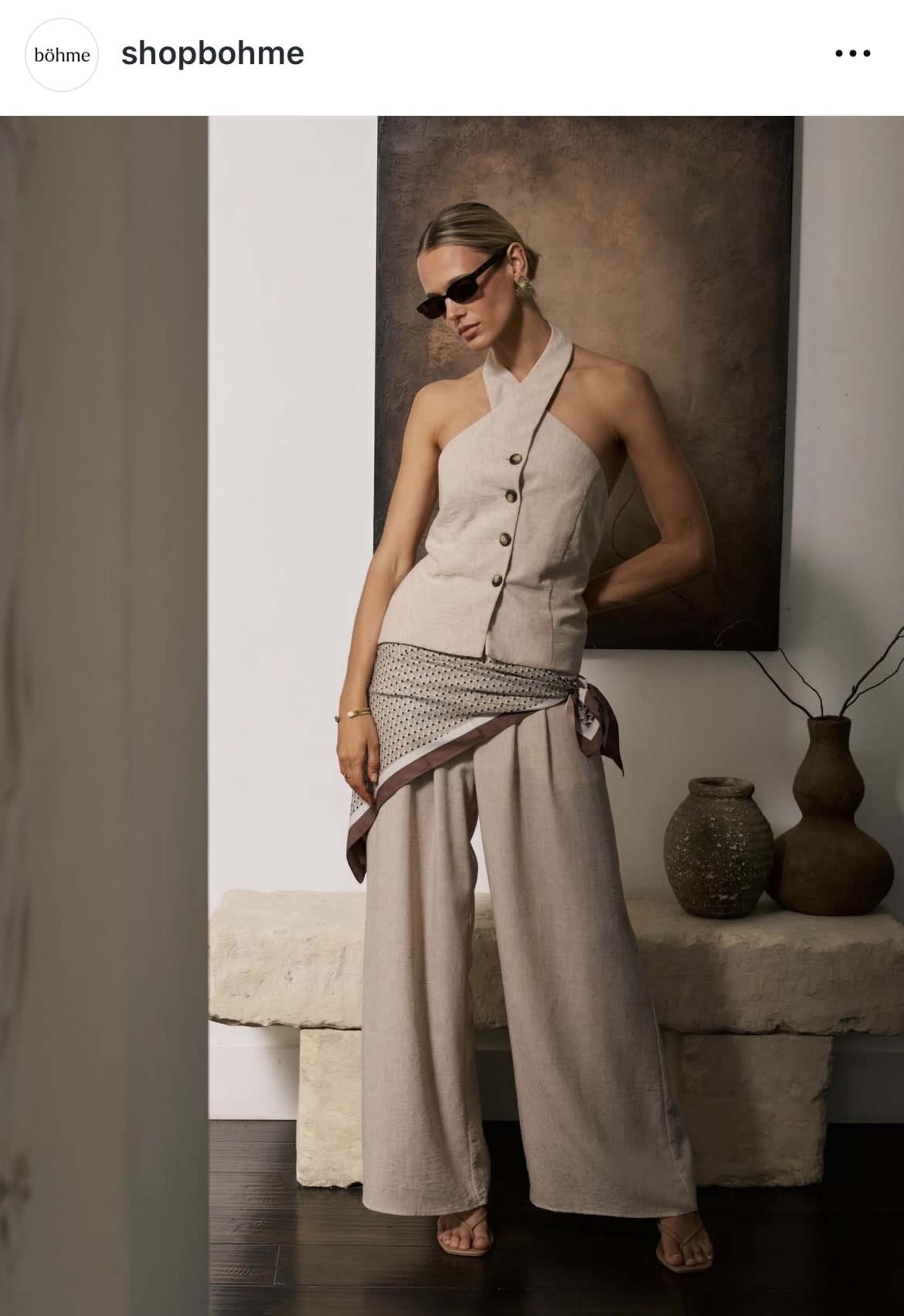 A woman poses in a stylish outfit with a one-shoulder top and wide-leg pants, accompanied by sunglasses and a scarf, against a minimalist backdrop with vases.