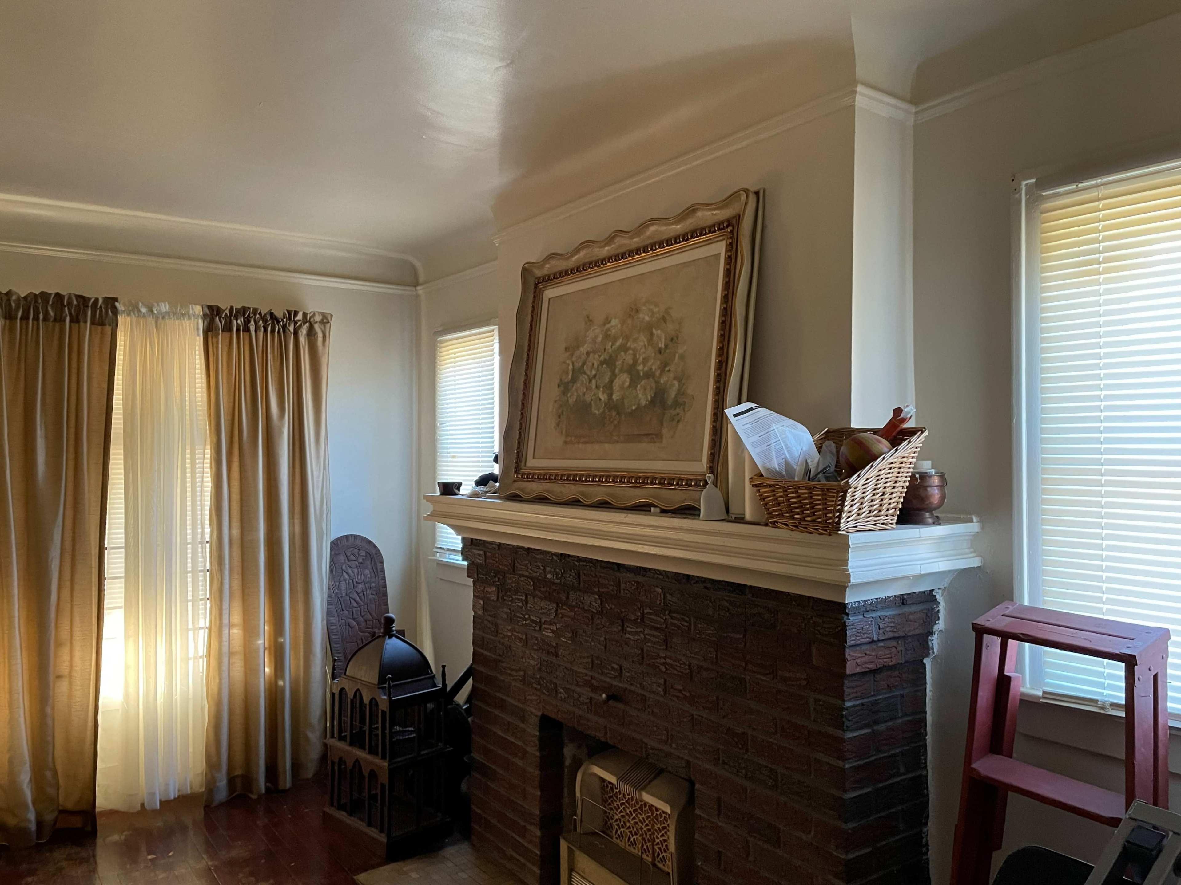 A living room with a fireplace, framed artwork above it, a basket on the mantle, and two windows with light curtains.