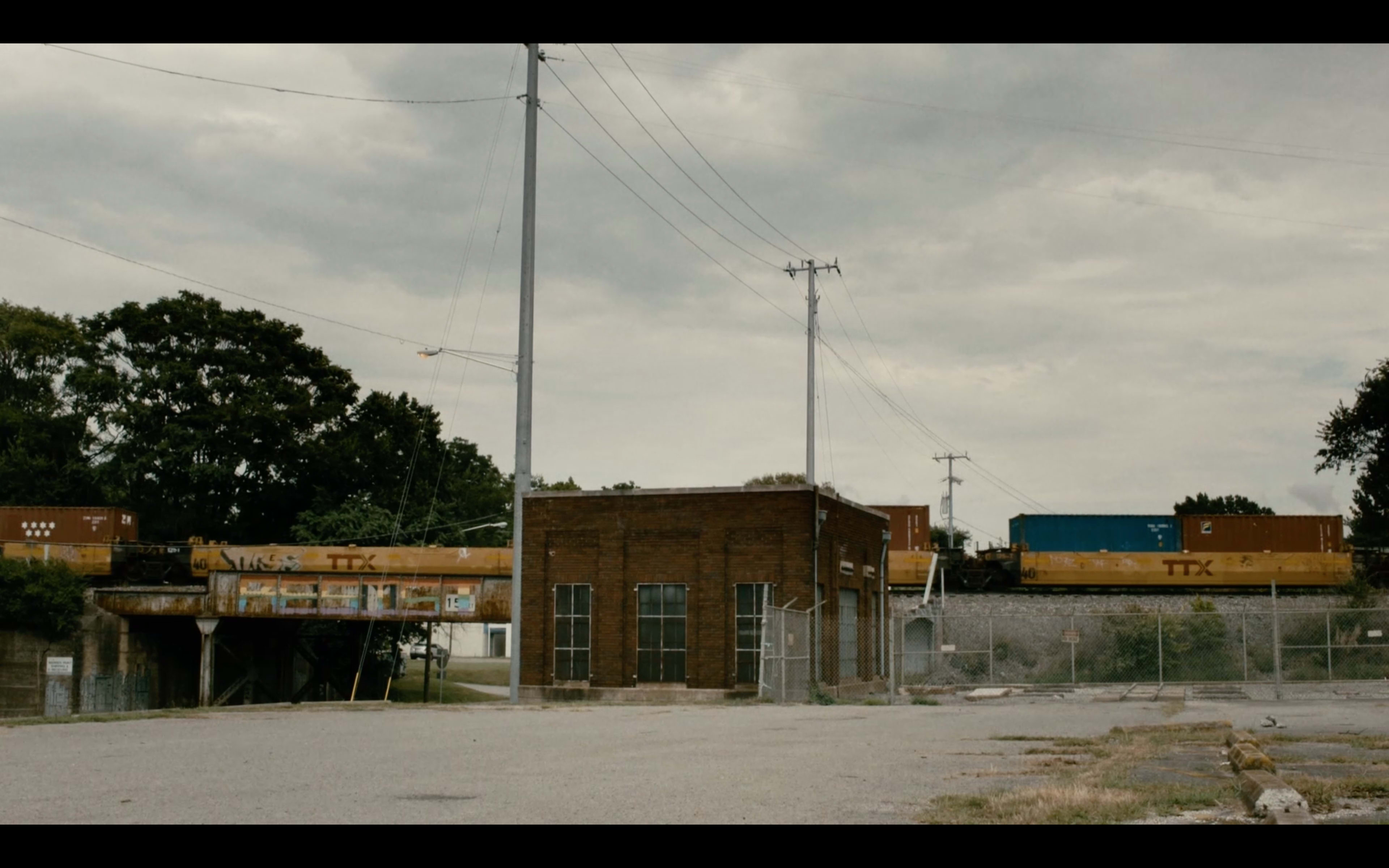 A line of freight train cars is parked beside an old brick building in an industrial area under a cloudy sky.