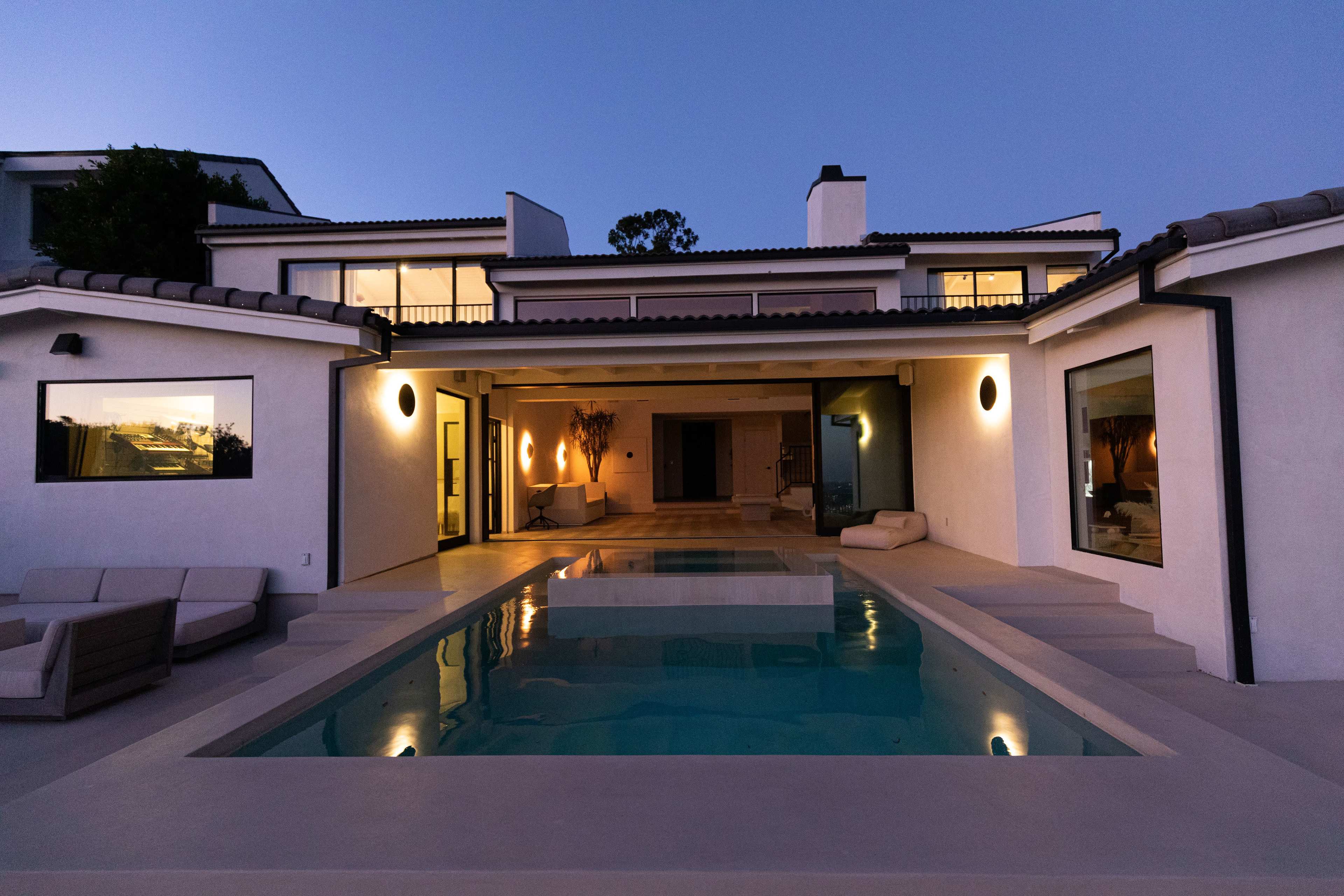 A modern white house with a swimming pool is illuminated at dusk, reflecting the surrounding architecture.