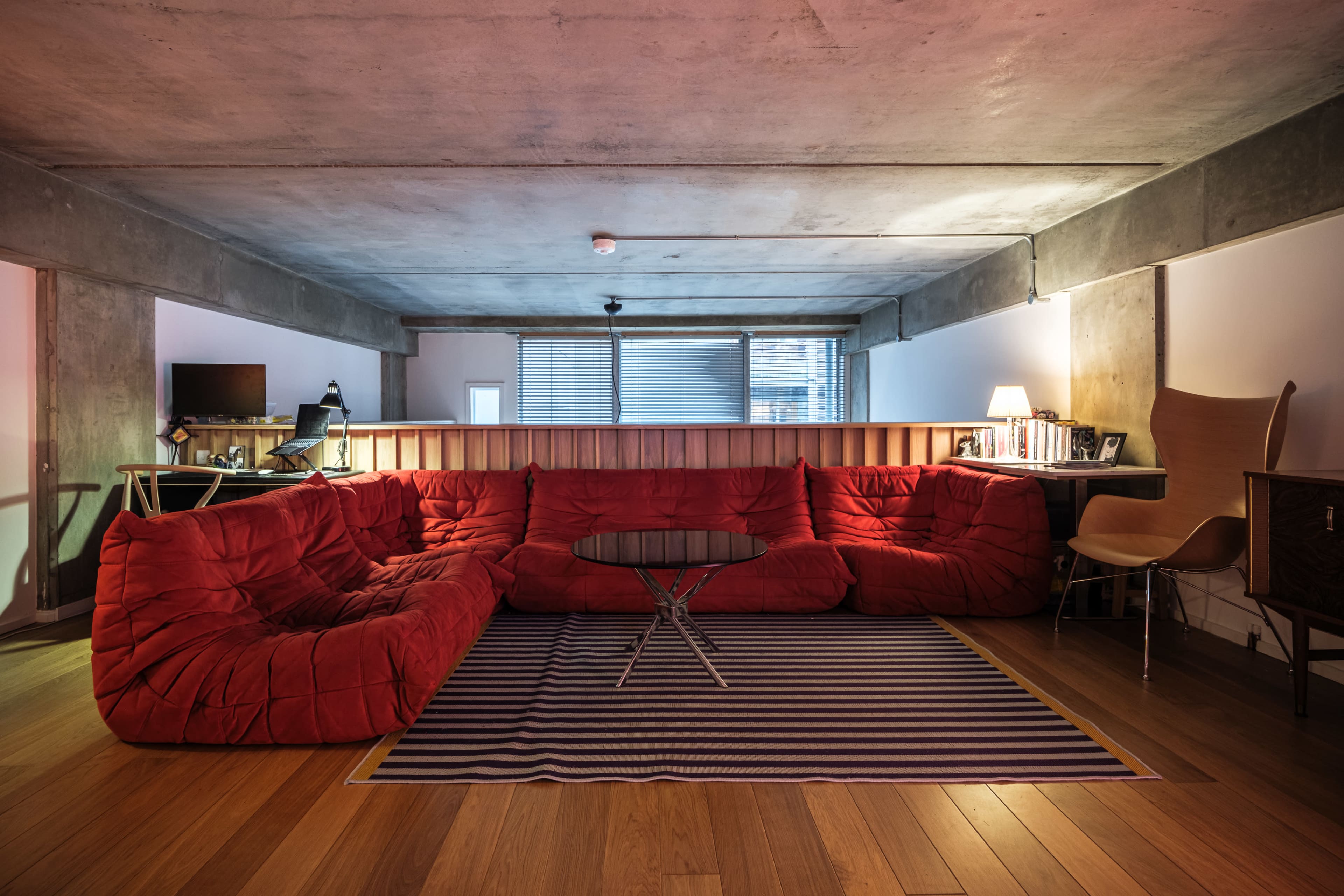 The image shows a spacious living room with a red, sectional sofa, a striped rug, a coffee table, and a wall of windows providing natural light.