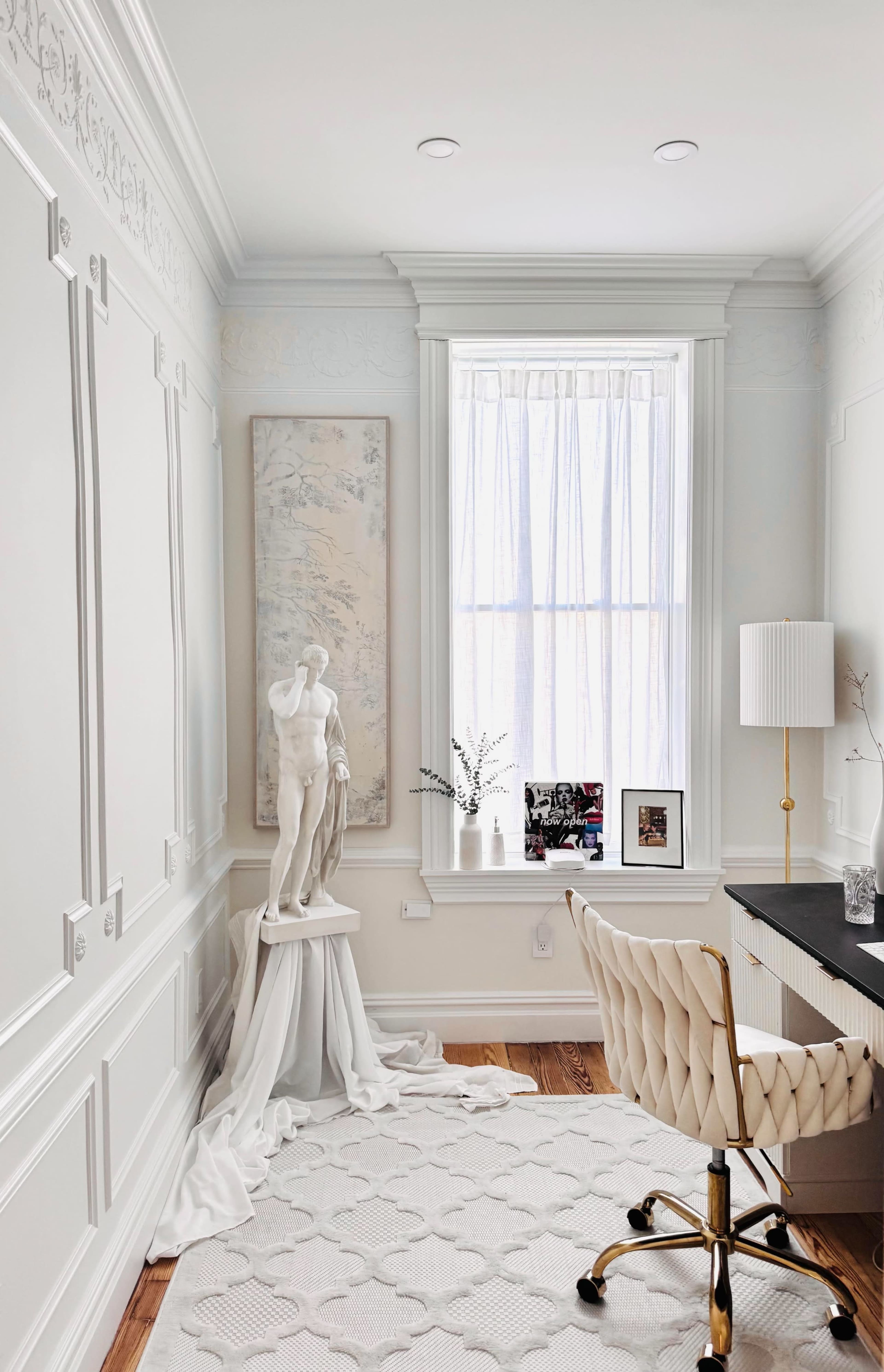 The image depicts a well-lit interior office space featuring a desk, a decorative statue on a pedestal, framed artwork on the wall, and a large window with sheer curtains.