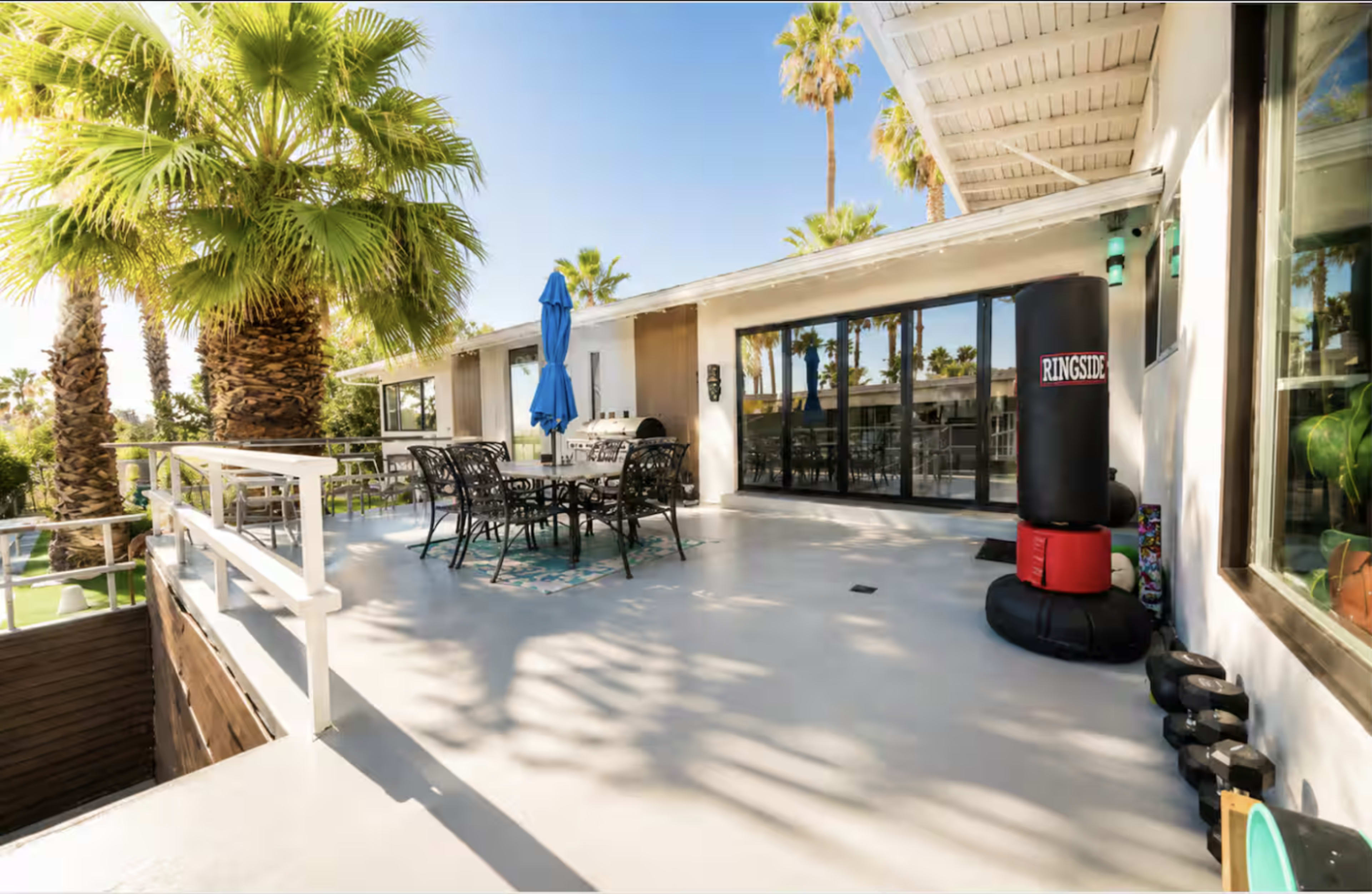 The image shows a spacious outdoor patio area surrounded by palm trees, featuring a table and chairs, a blue umbrella, and a boxing bag.