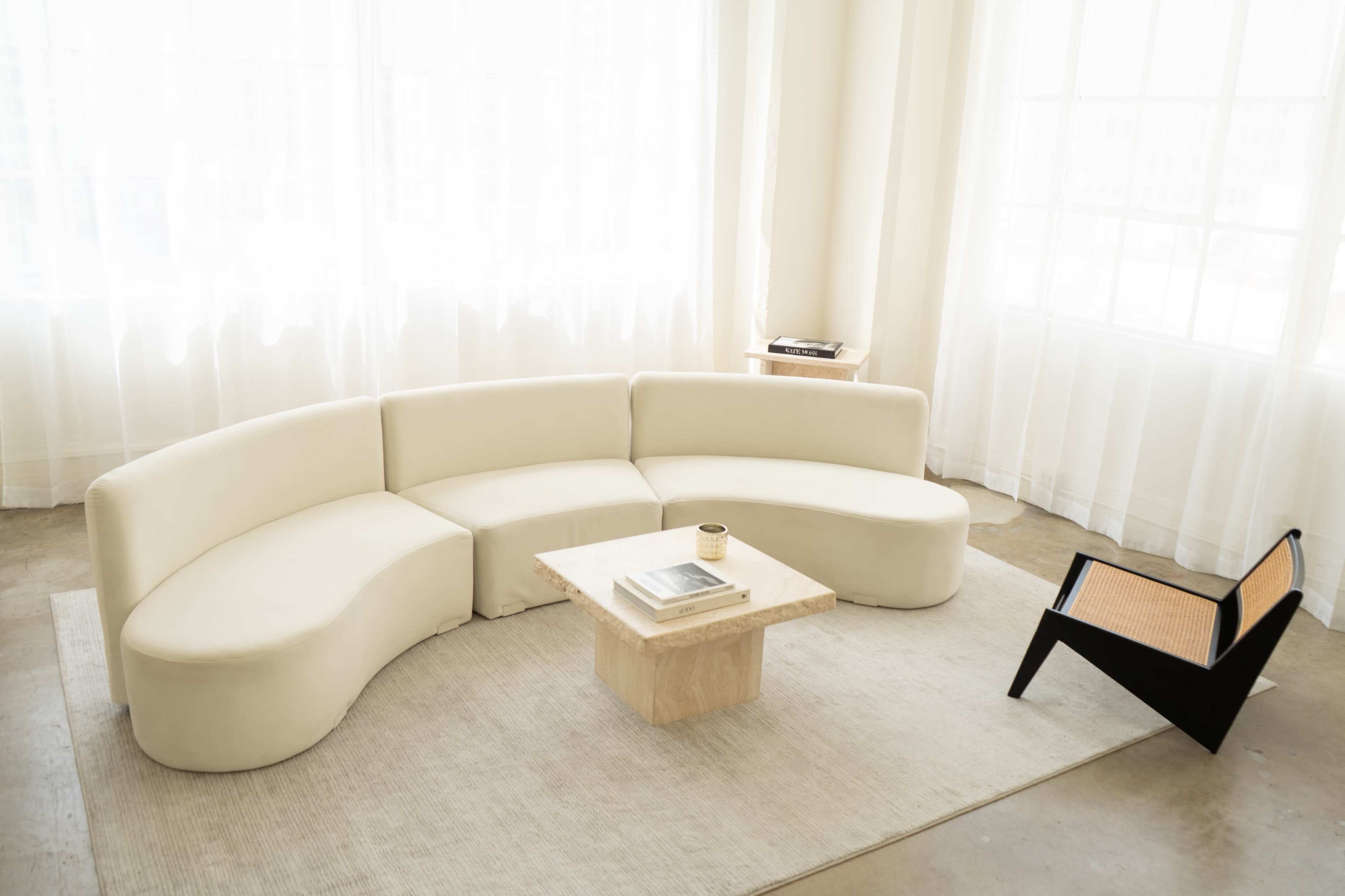 The scene features a modern, curved white sofa and a minimalist square coffee table on a light-colored rug beside a black accent chair, all set in a bright room with large windows and sheer curtains.
