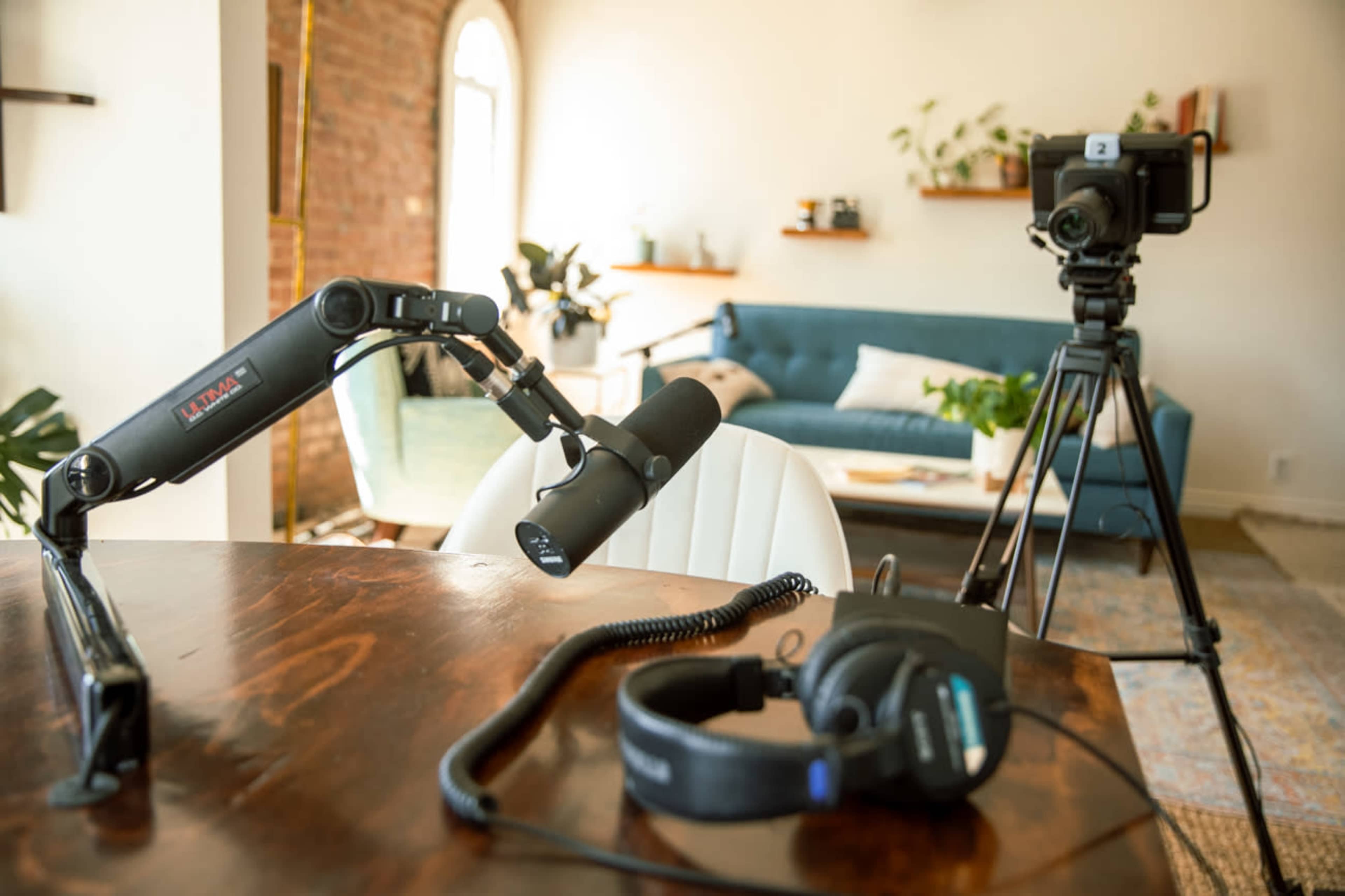 A at-home recording setup features a microphone on a boom arm, headphones on a table, and a camera positioned on a tripod near a sofa and plants.