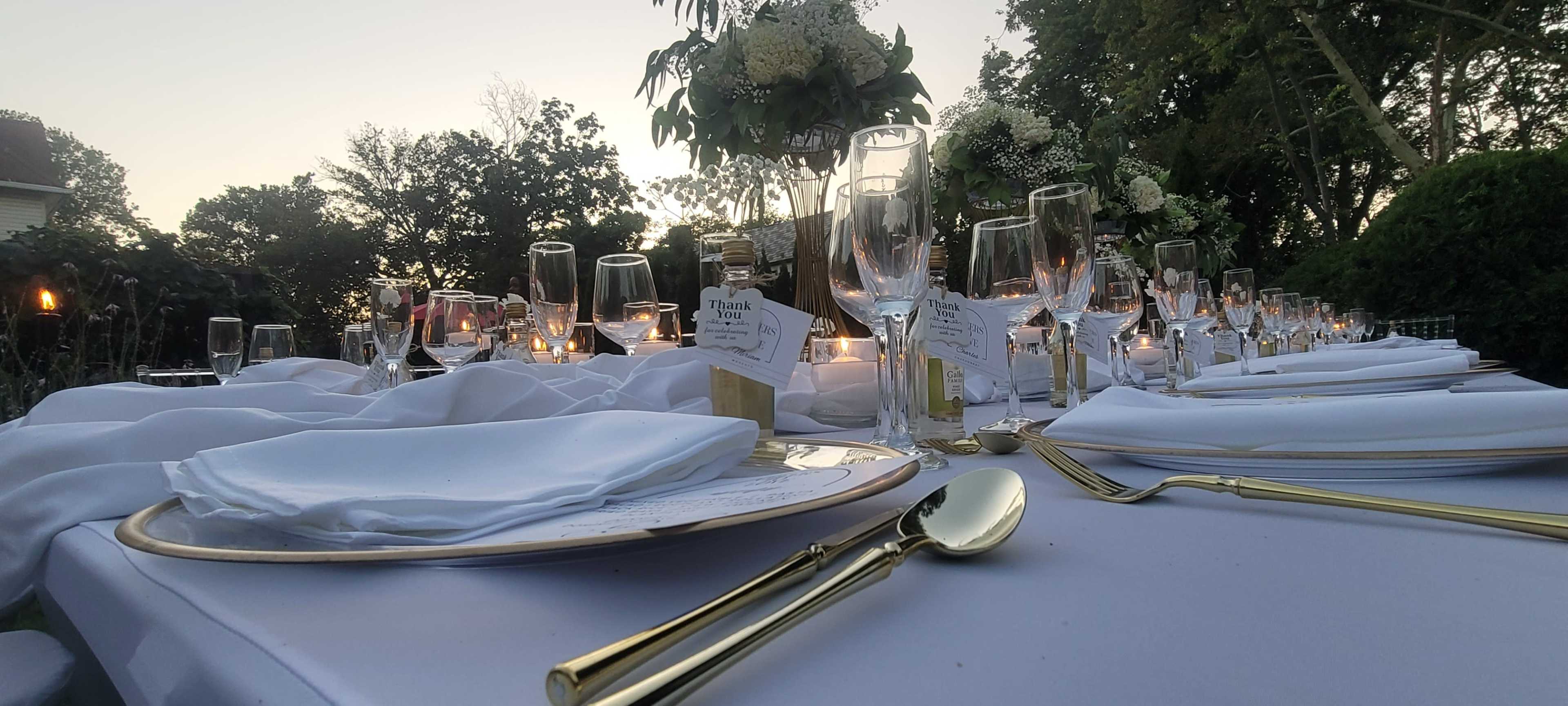 A long dining table is set outdoors with elegant place settings, glassware, and a floral centerpiece under a twilight sky.
