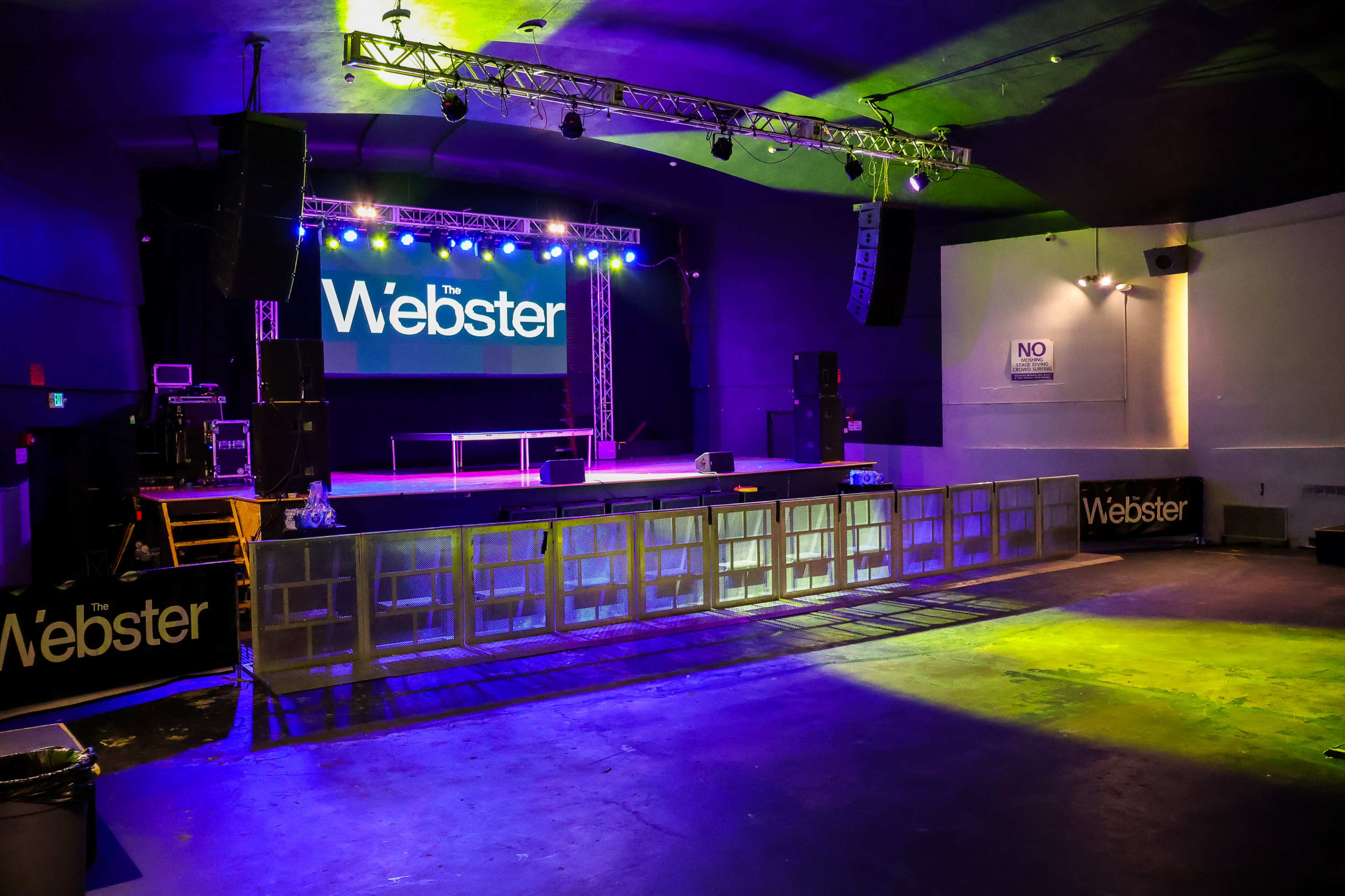 An empty concert stage at The Webster, with lighting set up and a large screen displaying the venue's name.