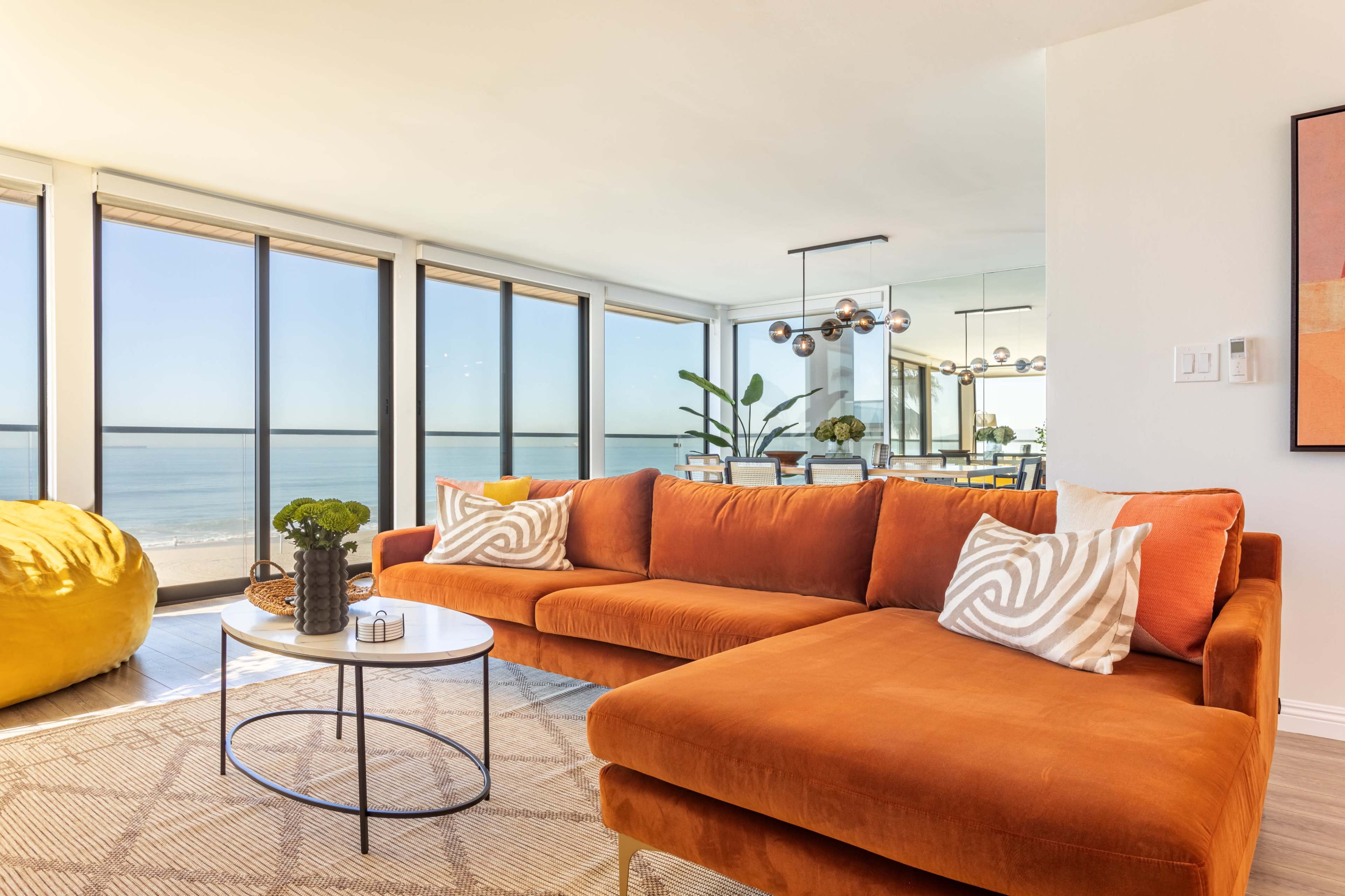 A modern living room features an orange sofa, a round coffee table, and large windows offering a view of the ocean.