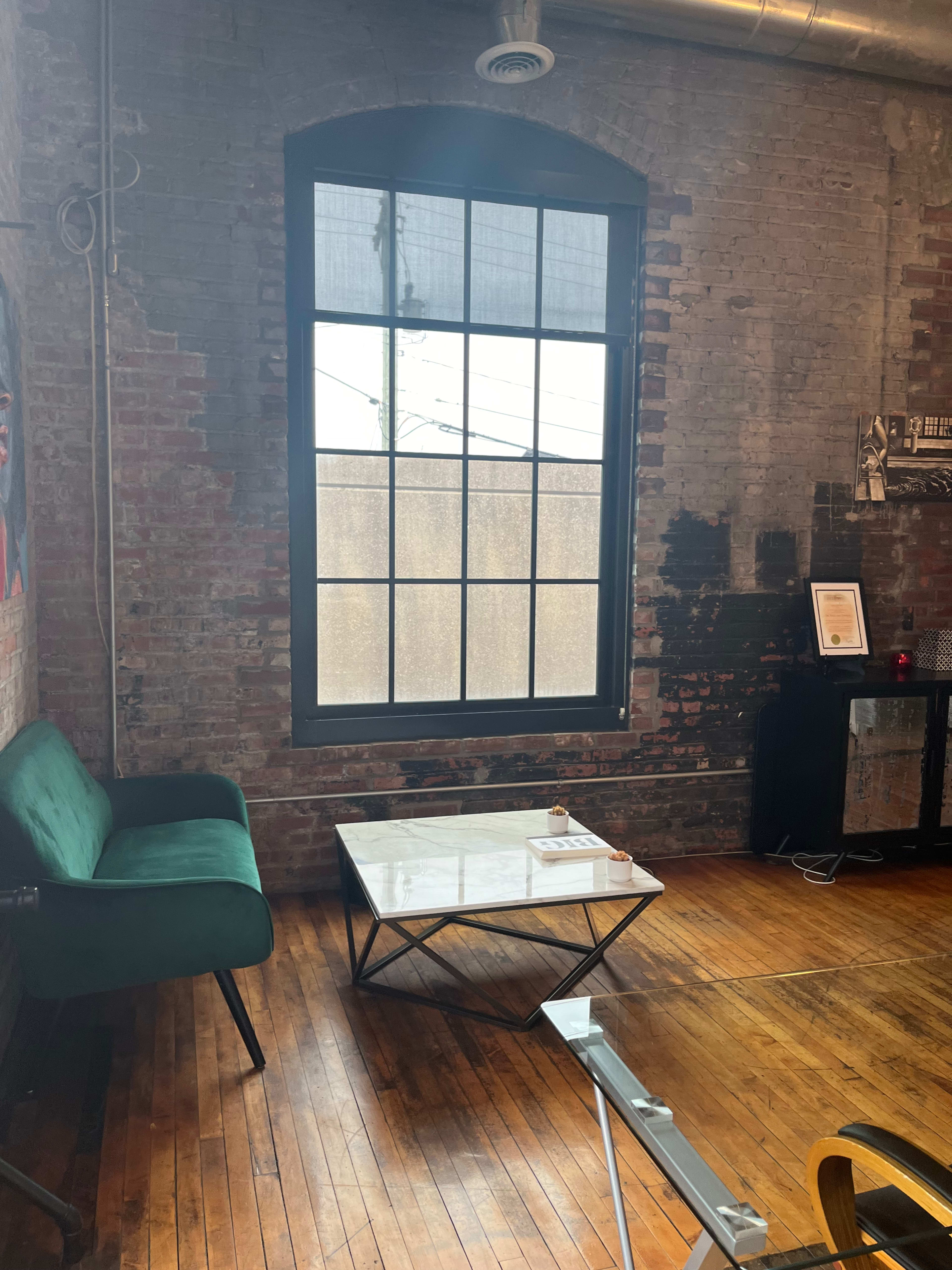 A room with exposed brick walls, a large window, a green sofa, a glass coffee table, and wooden flooring.