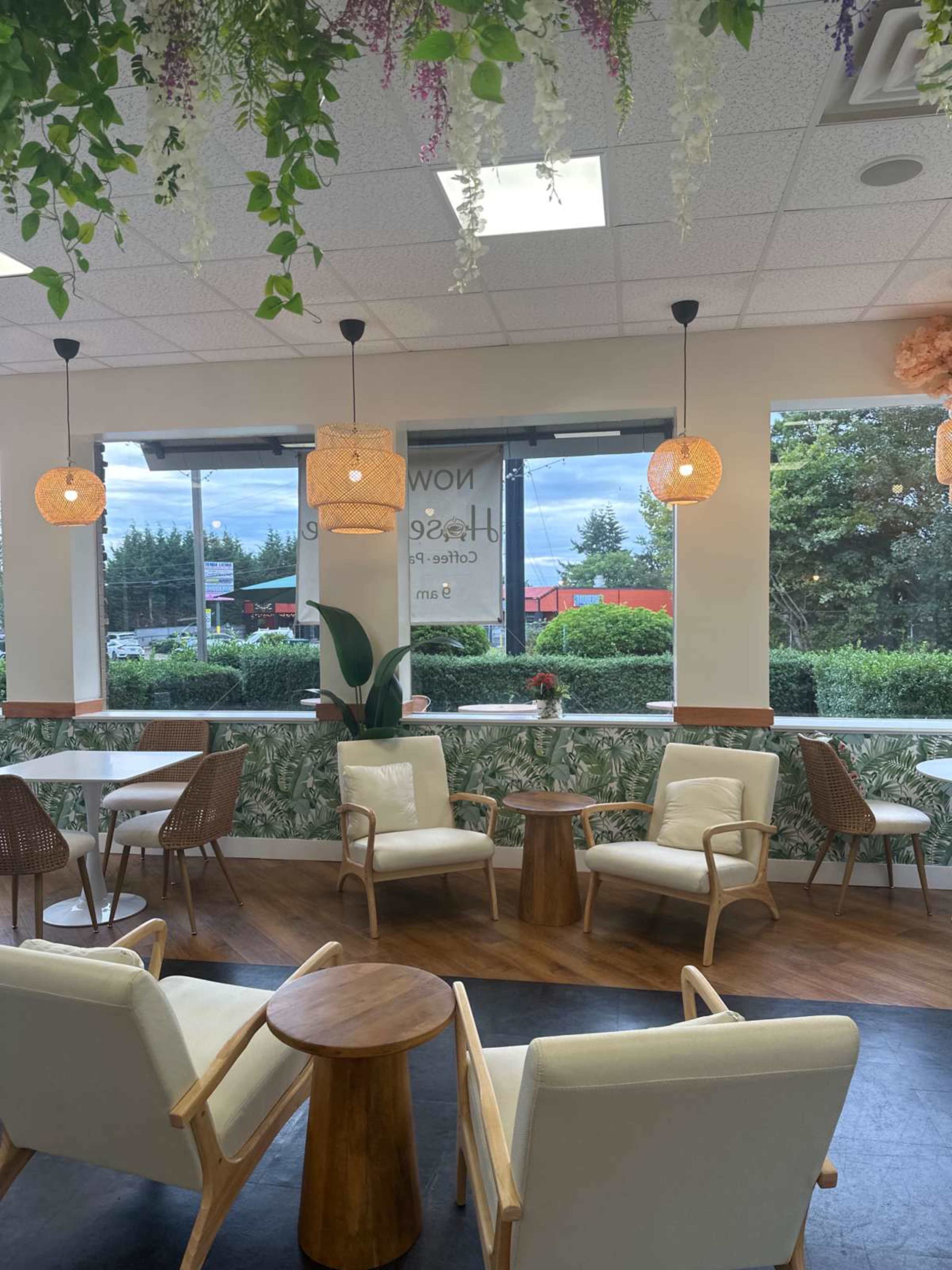 The image shows a cozy café interior with white chairs and wooden tables, large windows overlooking greenery, and decorative pendant lights.
