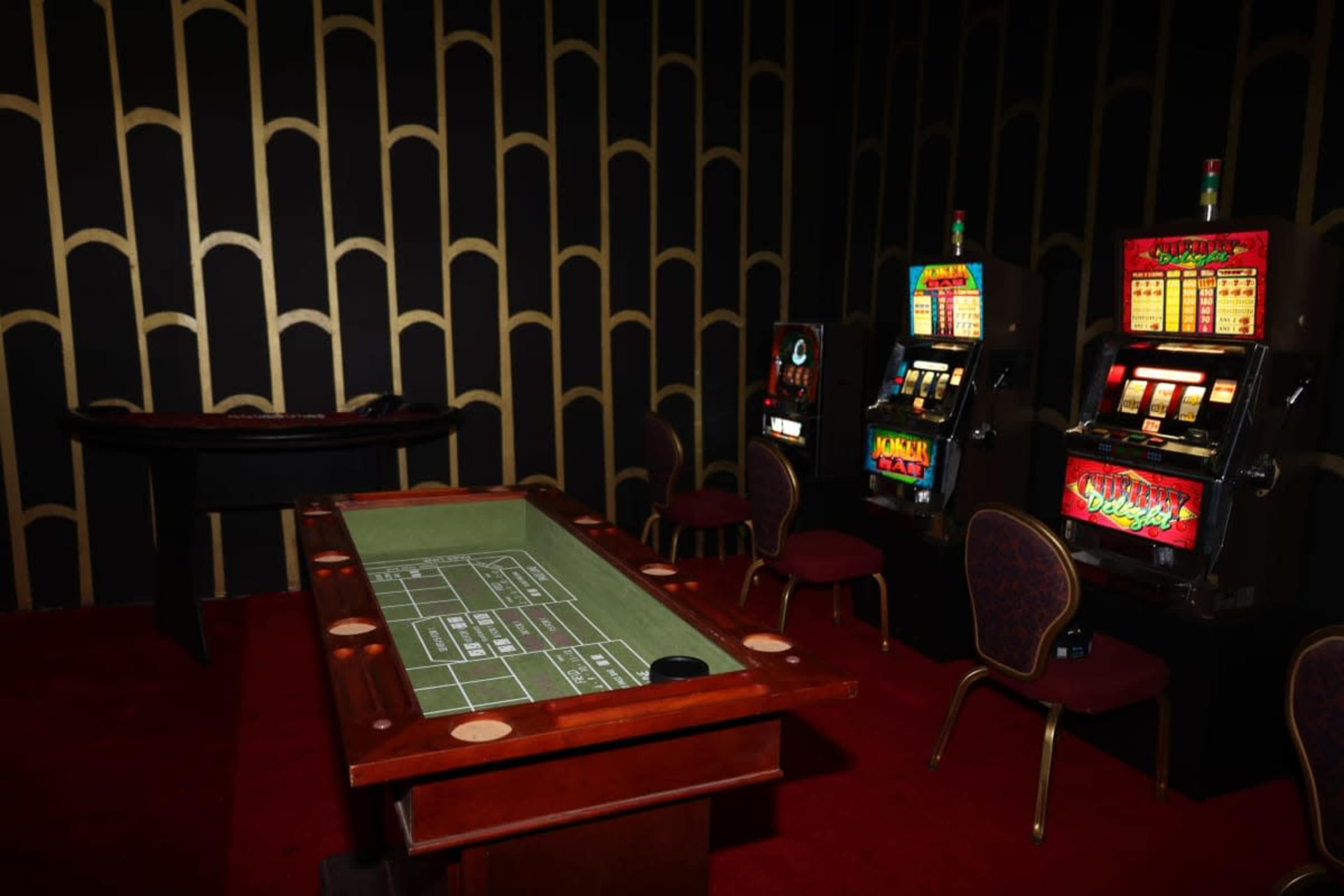 The image shows a dimly lit casino area with a green felt craps table and two slot machines against a decorative gold and black backdrop.
