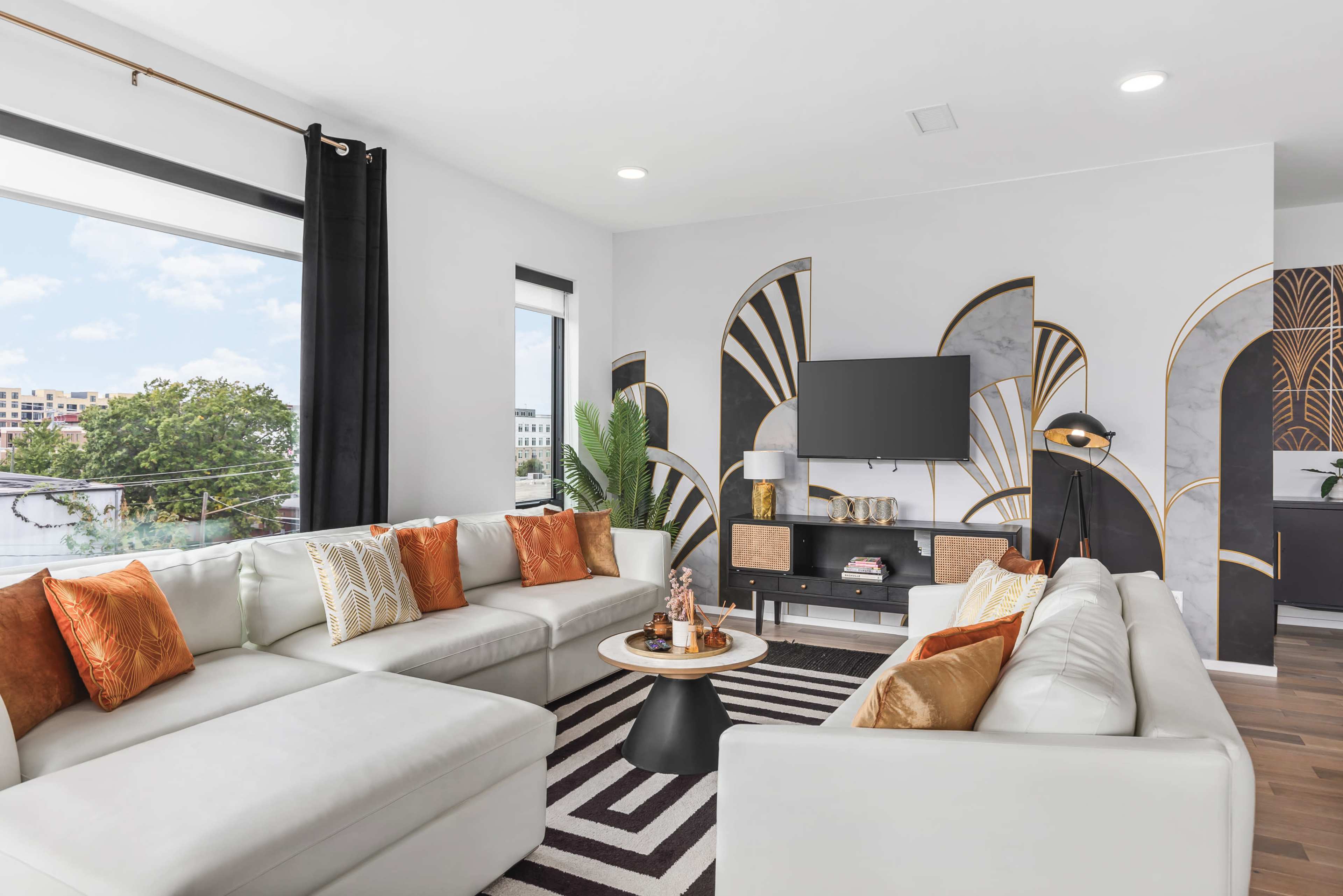 The image shows a modern living room with a white sectional sofa, decorative pillows, a round coffee table, and a patterned accent wall, featuring large windows and sleek furnishings.