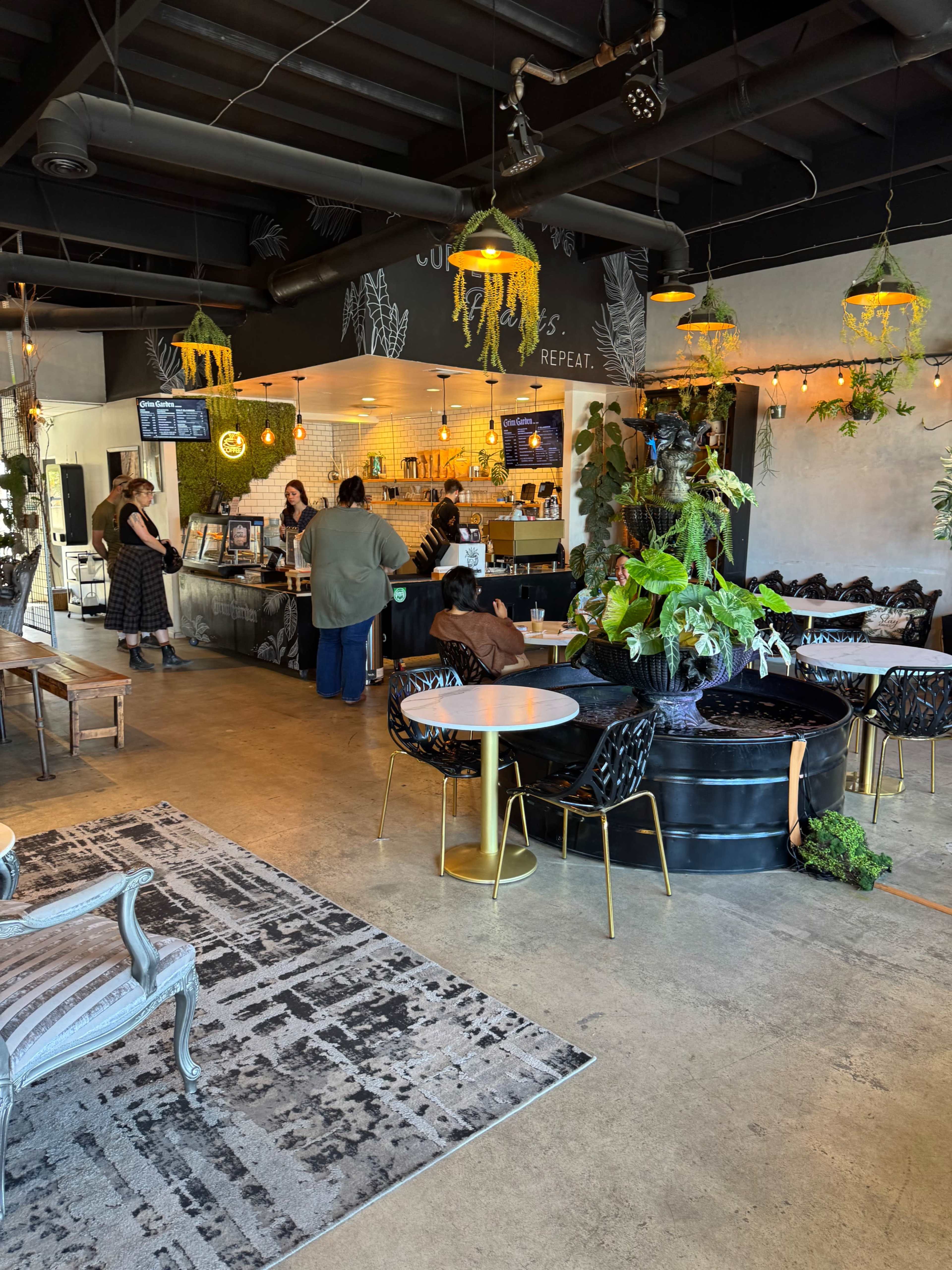 A café interior features a counter with staff serving customers, surrounded by comfortable seating areas and various plants hanging from the ceiling.