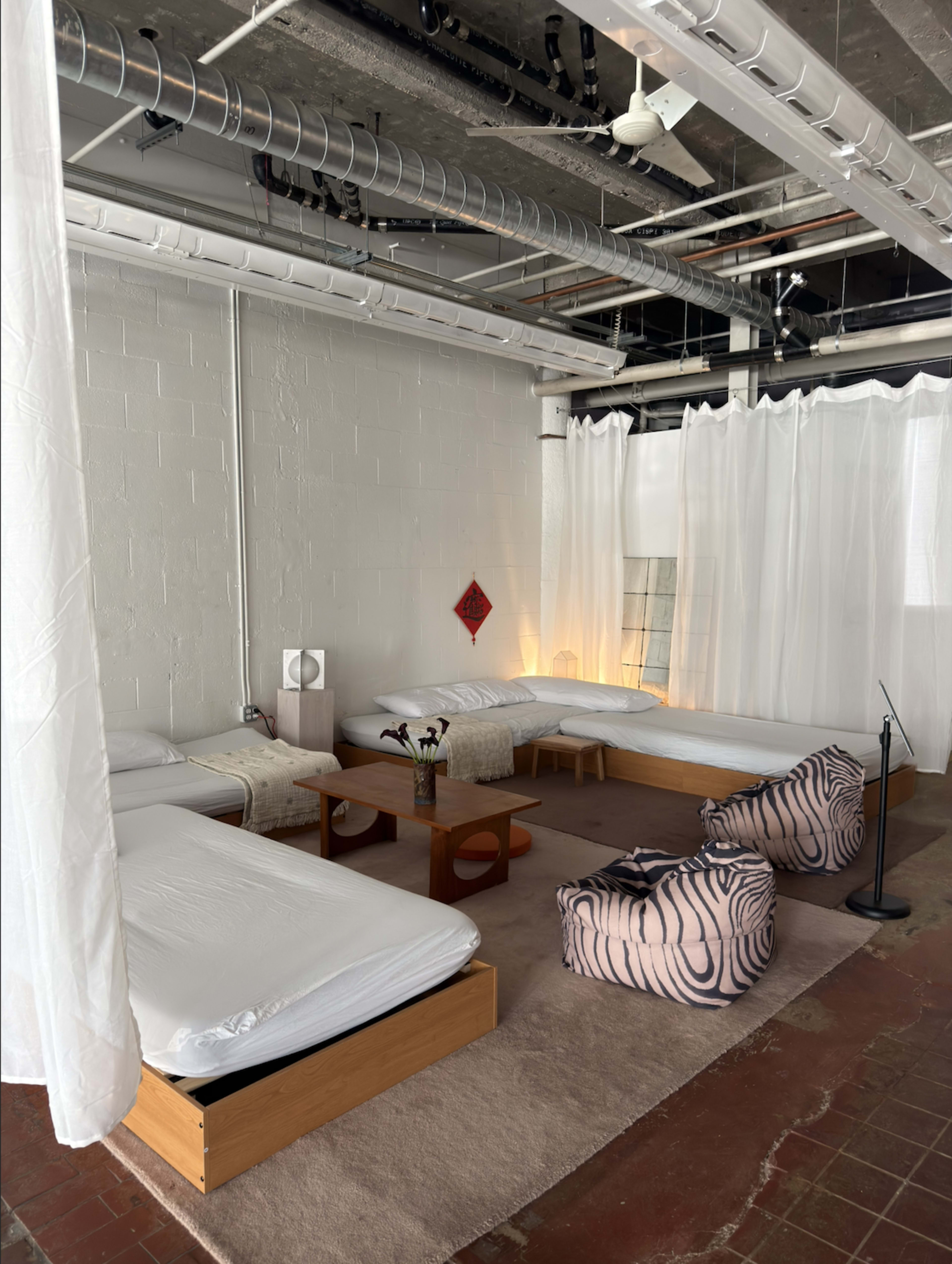 The image shows a spacious, minimalist room with several unmade beds, a low wooden table, and oversized floor cushions, all surrounded by sheer white curtains.